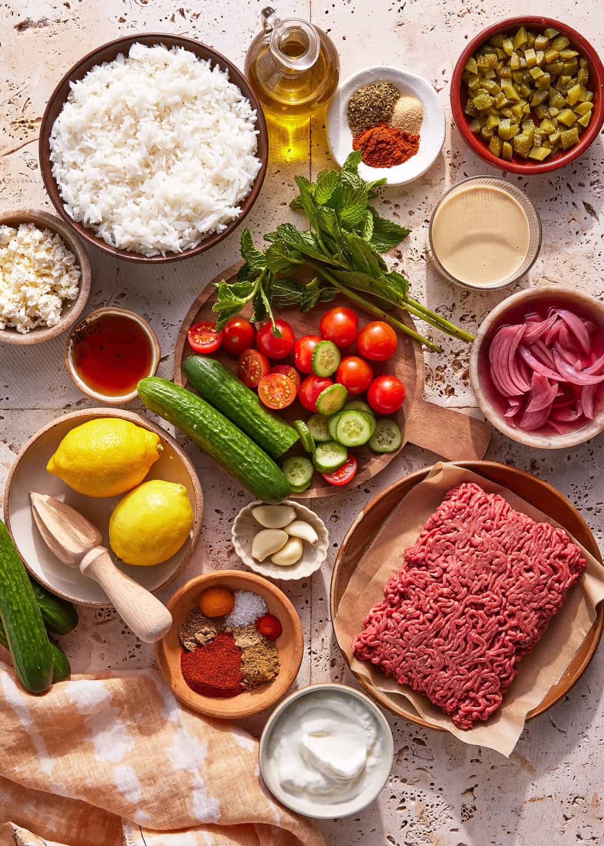 An assortment of ingredients on a light surface, including ground beef, cooked rice, cucumbers, cherry tomatoes, lemons, pickles, red onion, garlic, spices, olive oil, feta cheese, herbs, and a bowl of yogurt.