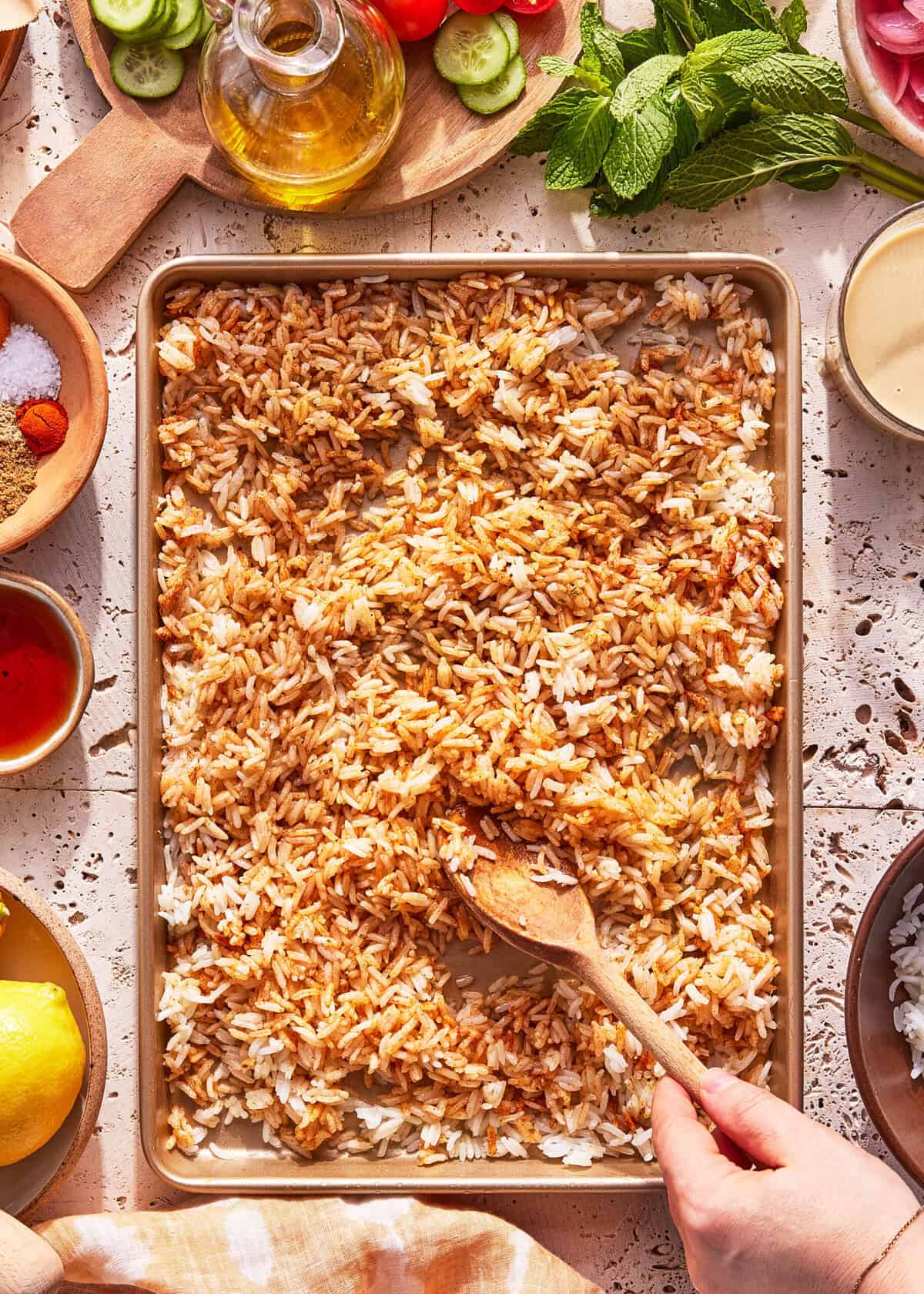 A hand stirs a mixture of cooked white and browned rice on a baking sheet with a wooden spoon. Surrounding the tray are bowls of spices, herbs, lemon, oil, and fresh vegetables on a textured surface.