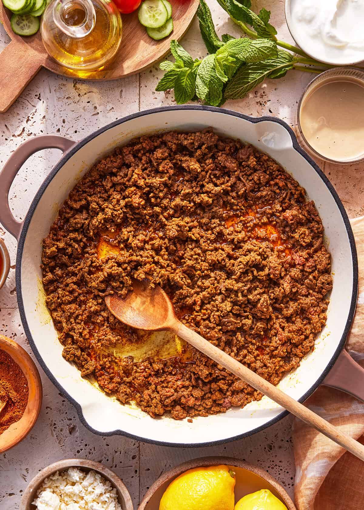 A skillet filled with cooked, seasoned ground beef, stirred with a wooden spoon. Surrounding the skillet are ingredients like lemon, feta cheese, fresh mint, olive oil, cucumber, tomatoes, and a bowl of white sauce.