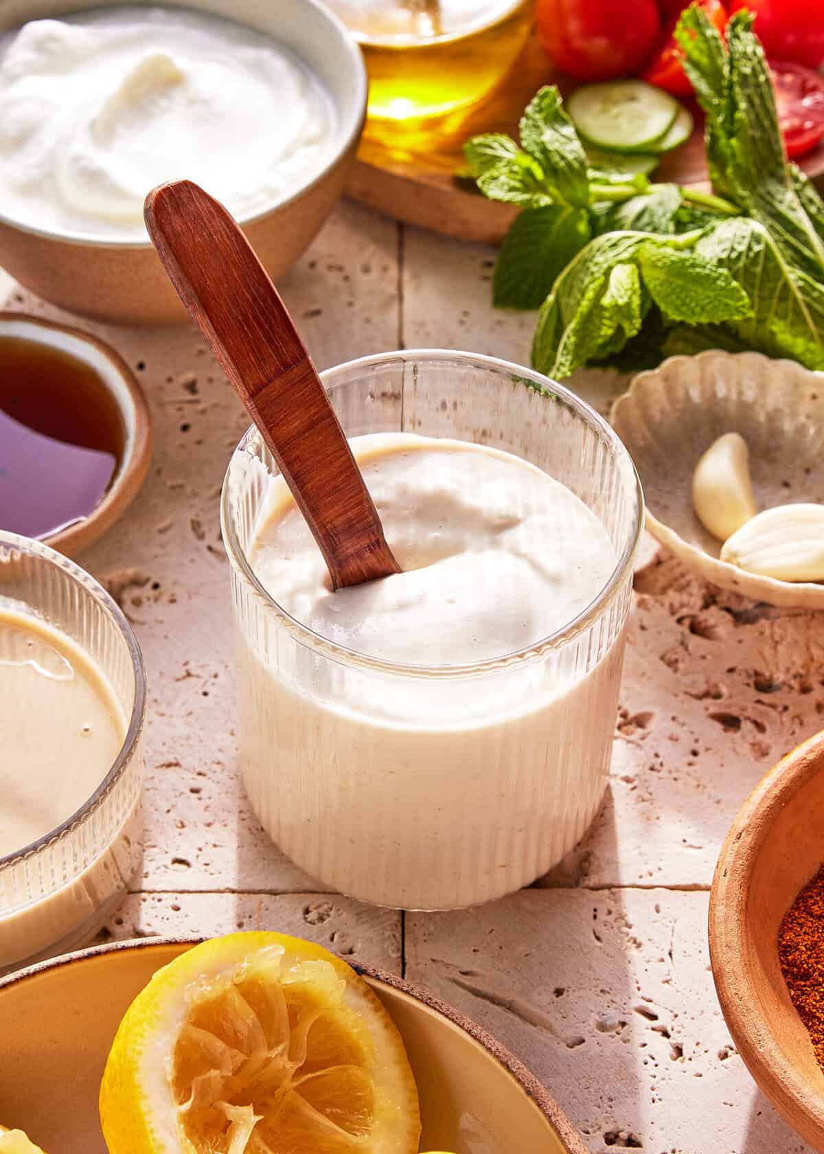A glass of creamy white sauce with a wooden spoon, surrounded by bowls of yogurt, honey, lemon, garlic, and fresh herbs on a textured beige surface.