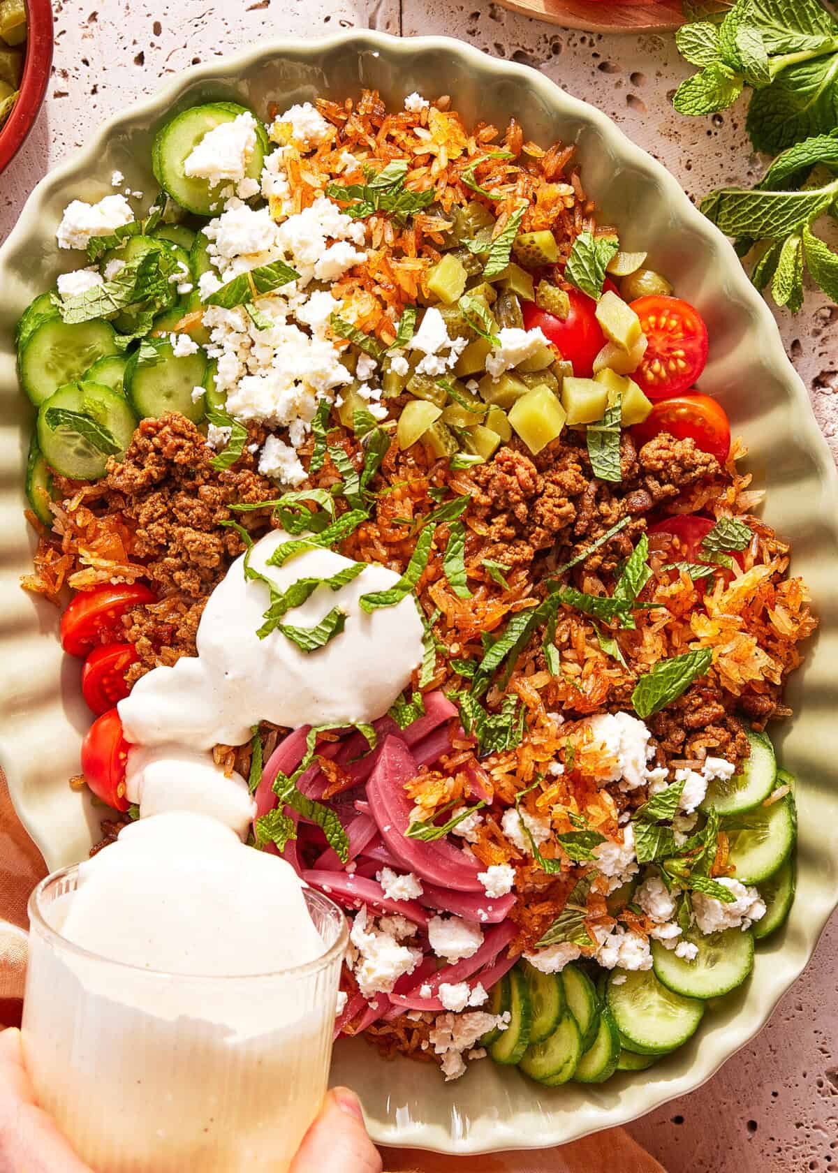 A hand pours creamy dressing over a platter with seasoned rice, ground meat, sliced cucumbers, cherry tomatoes, pickles, pickled onions, crumbled feta cheese, and fresh mint leaves.