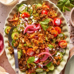 A large oval platter of salad with sliced cucumbers, cherry tomatoes, ground meat, rice, crumbled cheese, pickled onions, and fresh mint leaves, served with a spoon and surrounded by herbs and dressing.