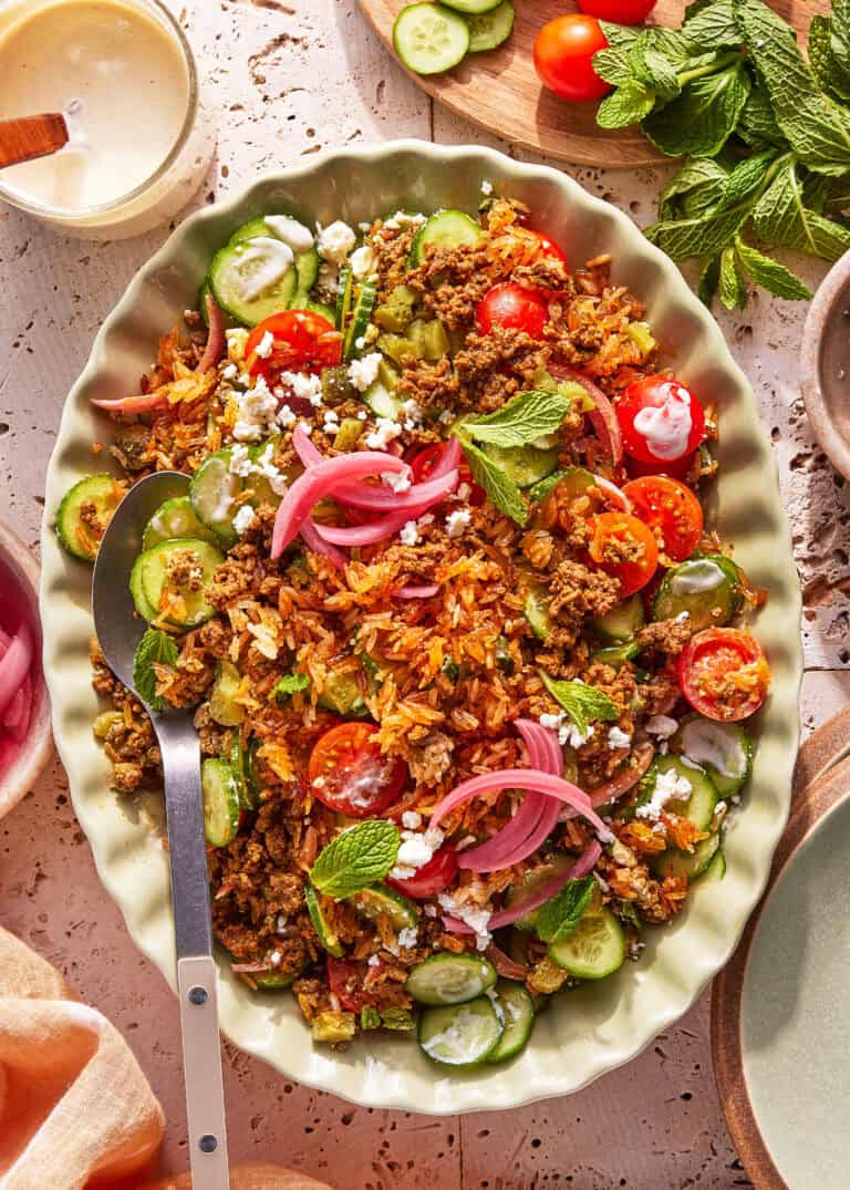 A large oval platter of salad with sliced cucumbers, cherry tomatoes, ground meat, rice, crumbled cheese, pickled onions, and fresh mint leaves, served with a spoon and surrounded by herbs and dressing.
