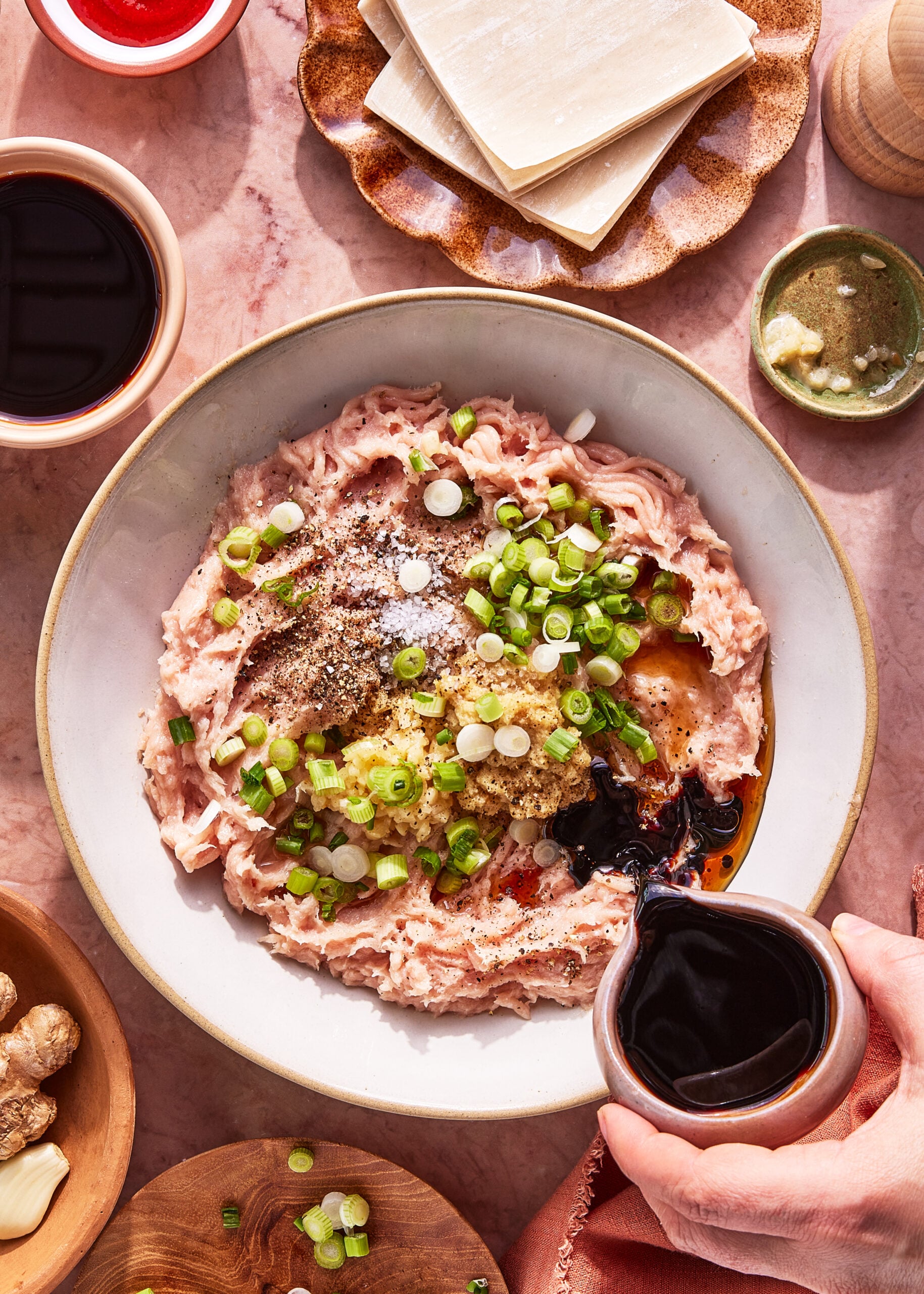 A bowl of ground meat topped with chopped green onions, minced garlic, and seasonings sits on a pink surface. A hand pours dark sauce into the bowl, surrounded by sauces, ginger, and a plate of square wonton wrappers.