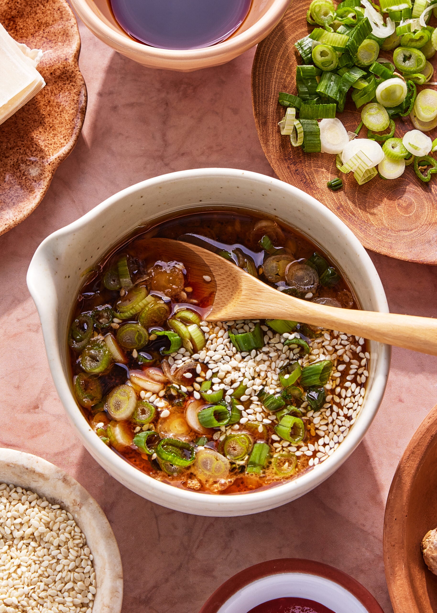A bowl of sauce with chopped green onions and sesame seeds, being mixed with a wooden spoon, surrounded by bowls of green onions, sesame seeds, and other ingredients on a pink surface.