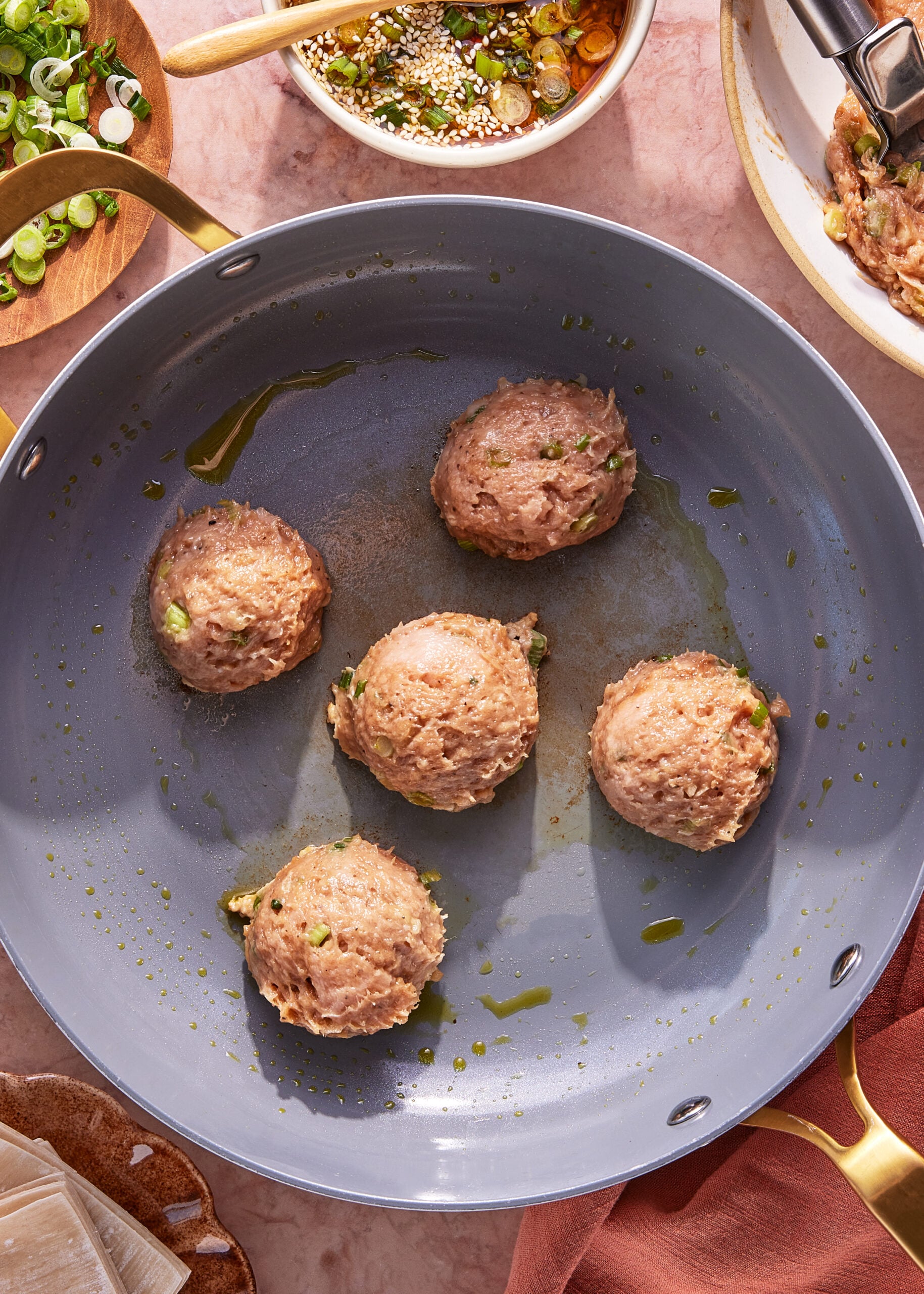Five raw meatballs with chopped green onions are arranged in a large, nonstick skillet with a light coating of oil. Surrounding the pan are bowls with chopped scallions, sauce, and a plate with stacked wonton wrappers.