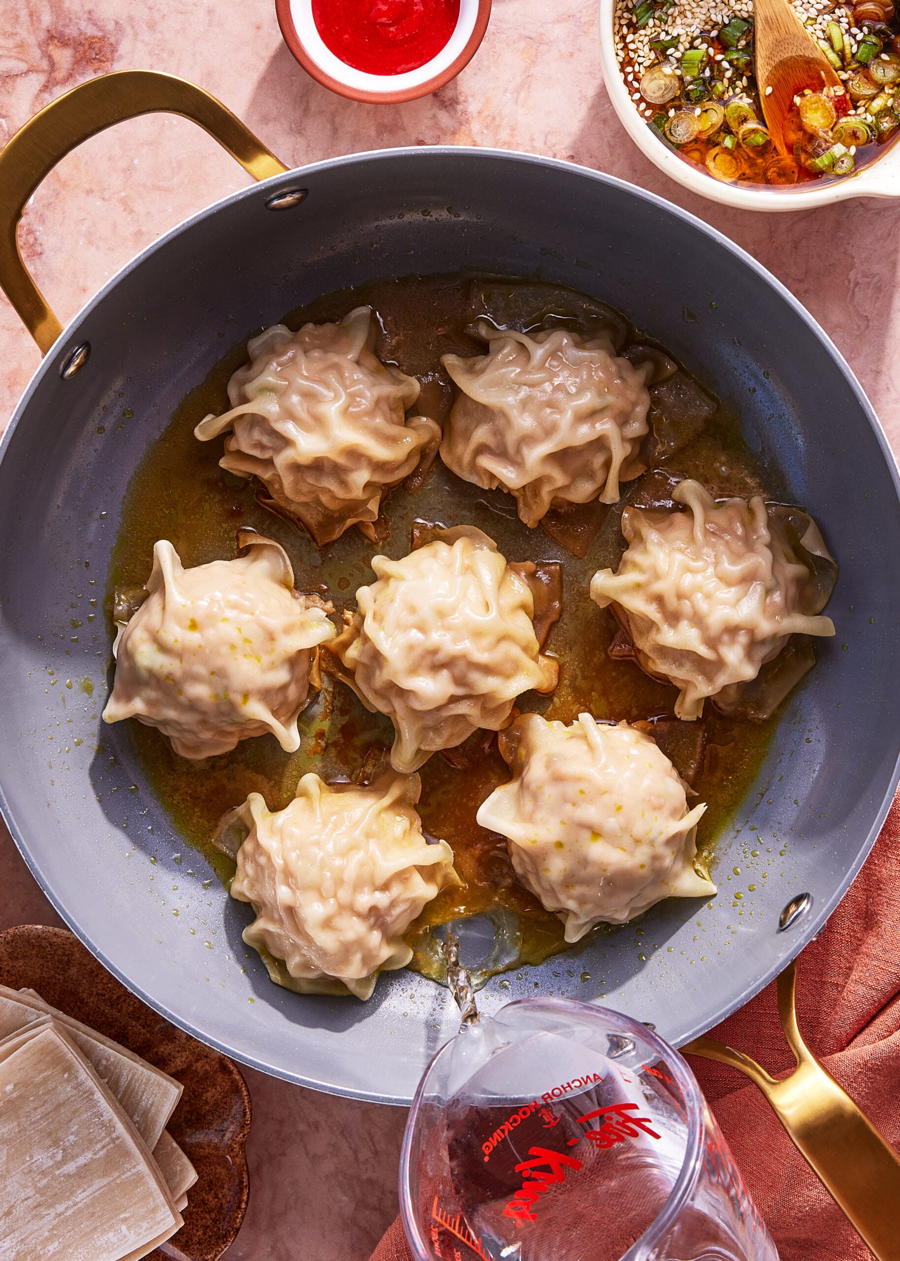 Overhead view of a pan with seven steamed dumplings in broth, surrounded by dipping sauces, wonton wrappers, and a measuring cup pouring liquid into the pan.