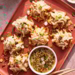 A red plate with seven steamed dumplings topped with chopped green onions and chili oil, served with a bowl of dipping sauce. Chopsticks and extra sauce bowls are nearby.