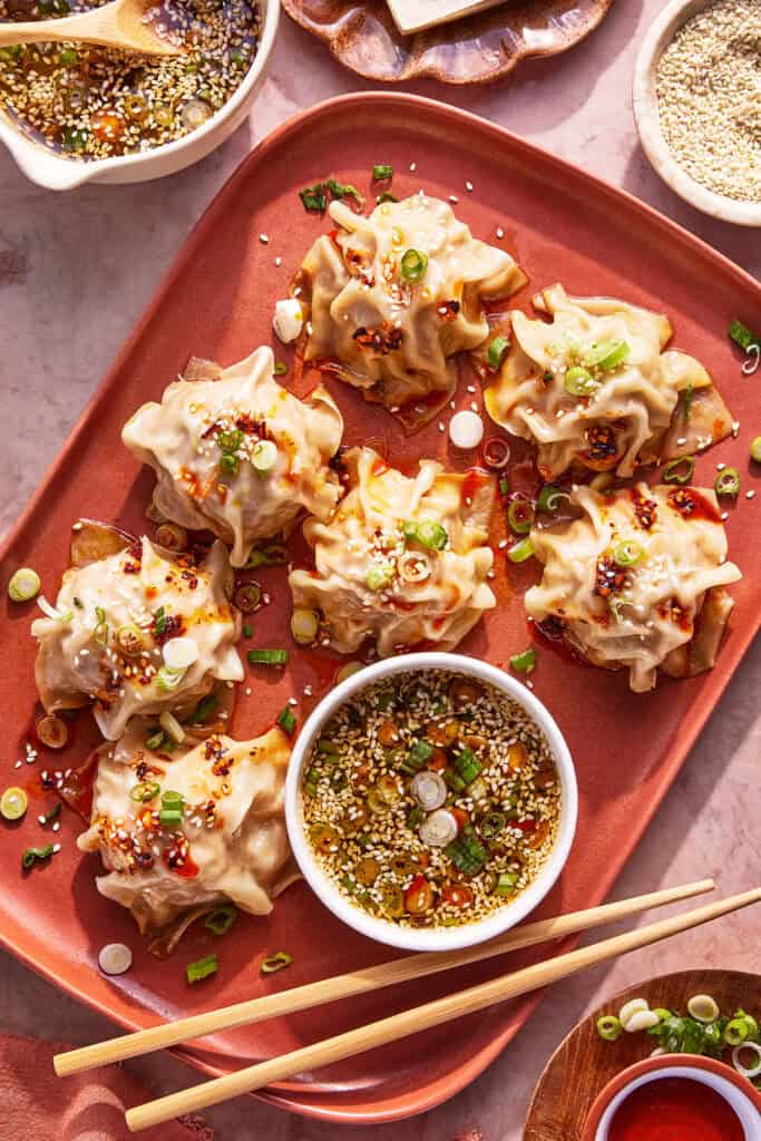 A red plate with seven steamed dumplings topped with chopped green onions and chili oil, served with a bowl of dipping sauce. Chopsticks and extra sauce bowls are nearby.