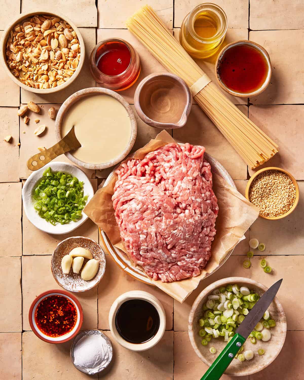 Ingredients for an Asian-inspired noodle dish arranged on a tiled surface: ground meat, dry spaghetti, chopped scallions, garlic cloves, sesame seeds, peanuts, sauces, oil, chili flakes, and a green-handled knife.