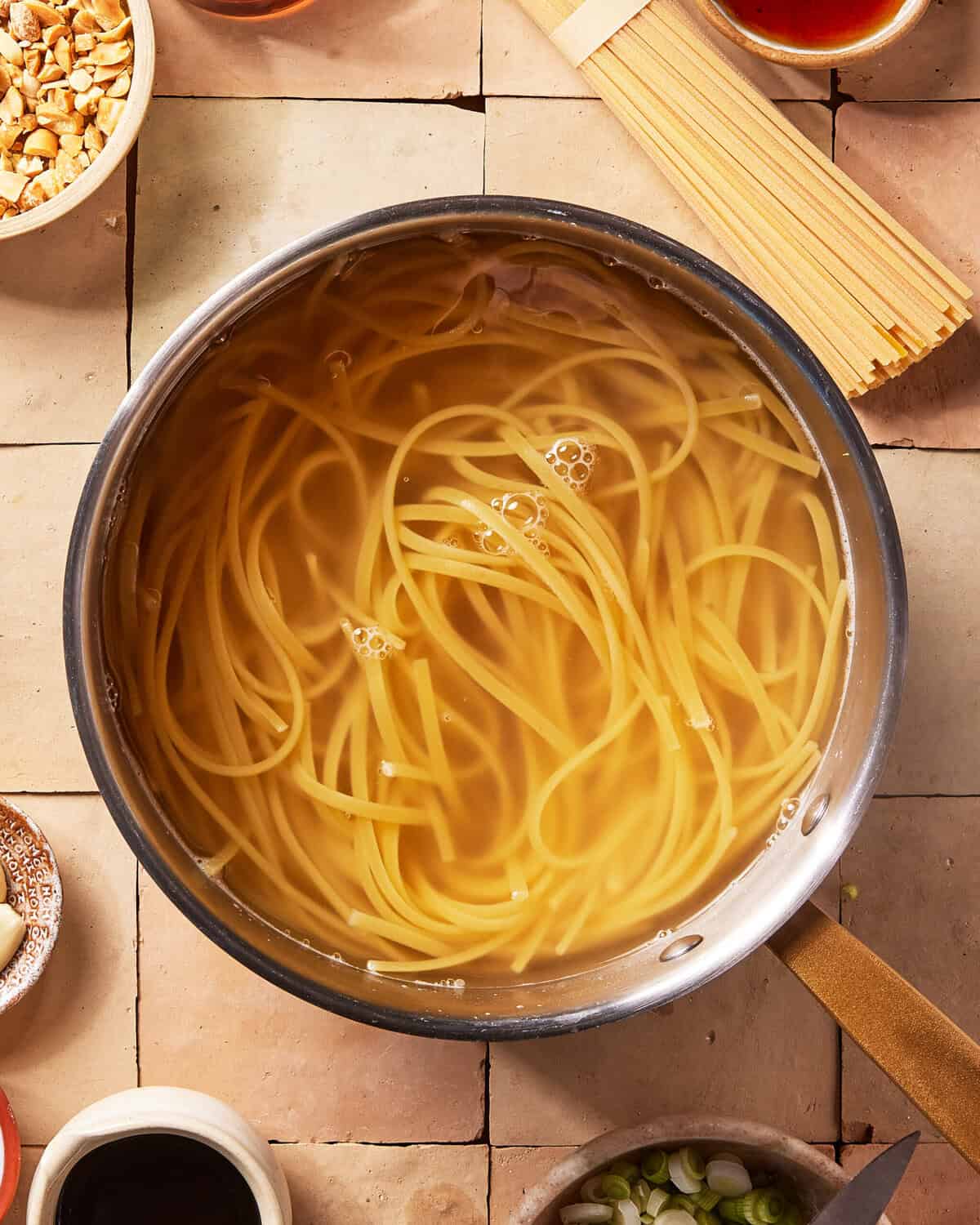 A pot of uncooked spaghetti noodles soaking in water, surrounded by small bowls of chopped nuts, green onions, and sauces on a tiled surface.