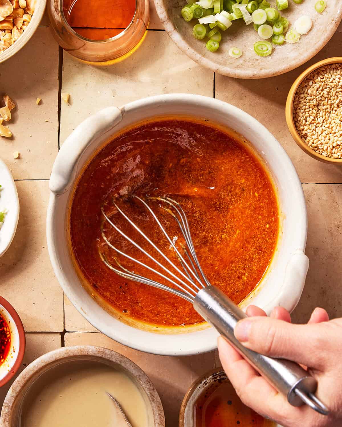 A hand uses a whisk to mix a reddish-brown sauce in a white bowl, surrounded by small bowls of sliced green onions, sesame seeds, nuts, sauces, and oil on a tiled surface.