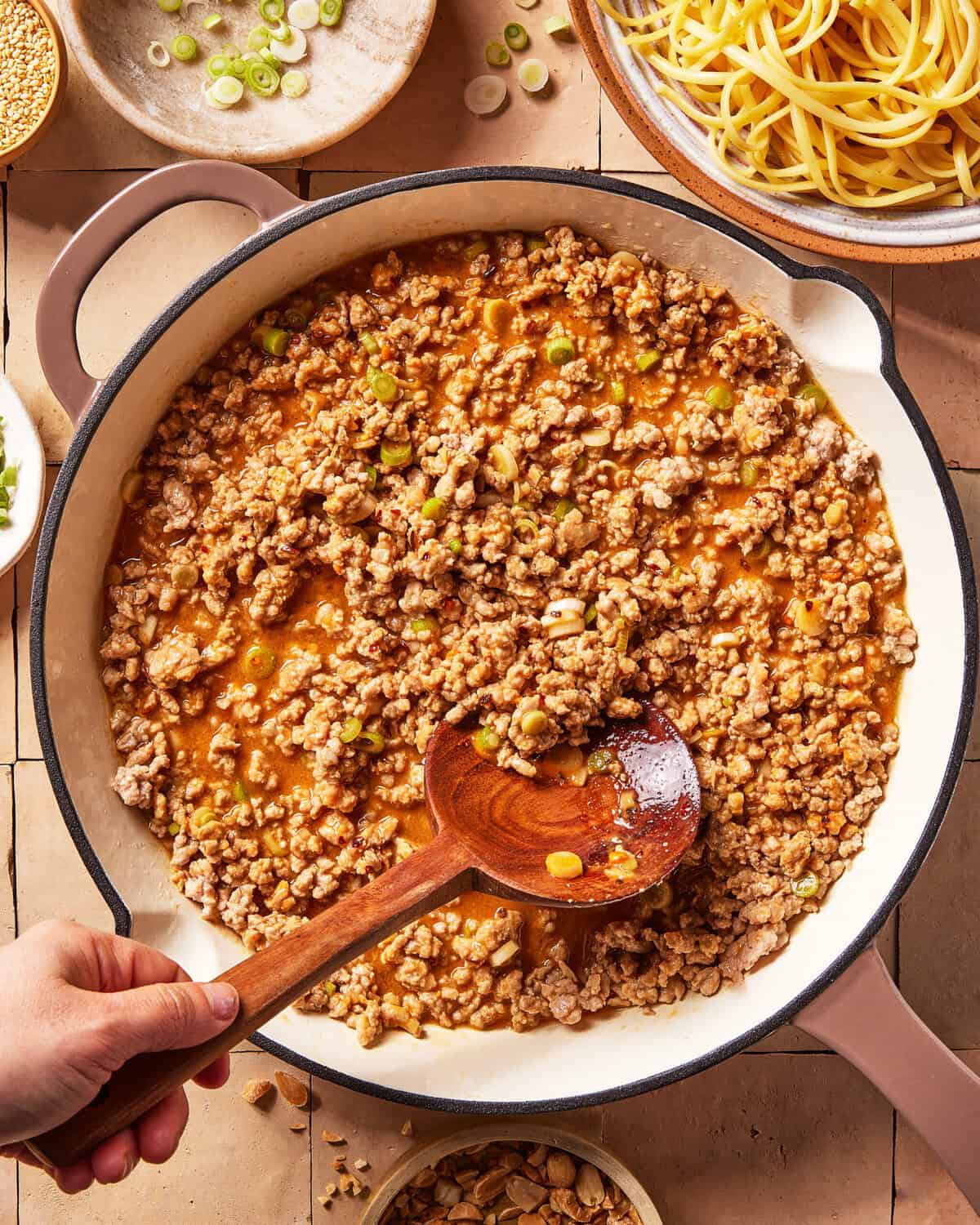 A hand stirs a pan of cooked ground meat in a savory sauce with chopped green onions. Surrounding the pan are bowls of uncooked noodles, peanuts, and sliced scallions on a tiled countertop.