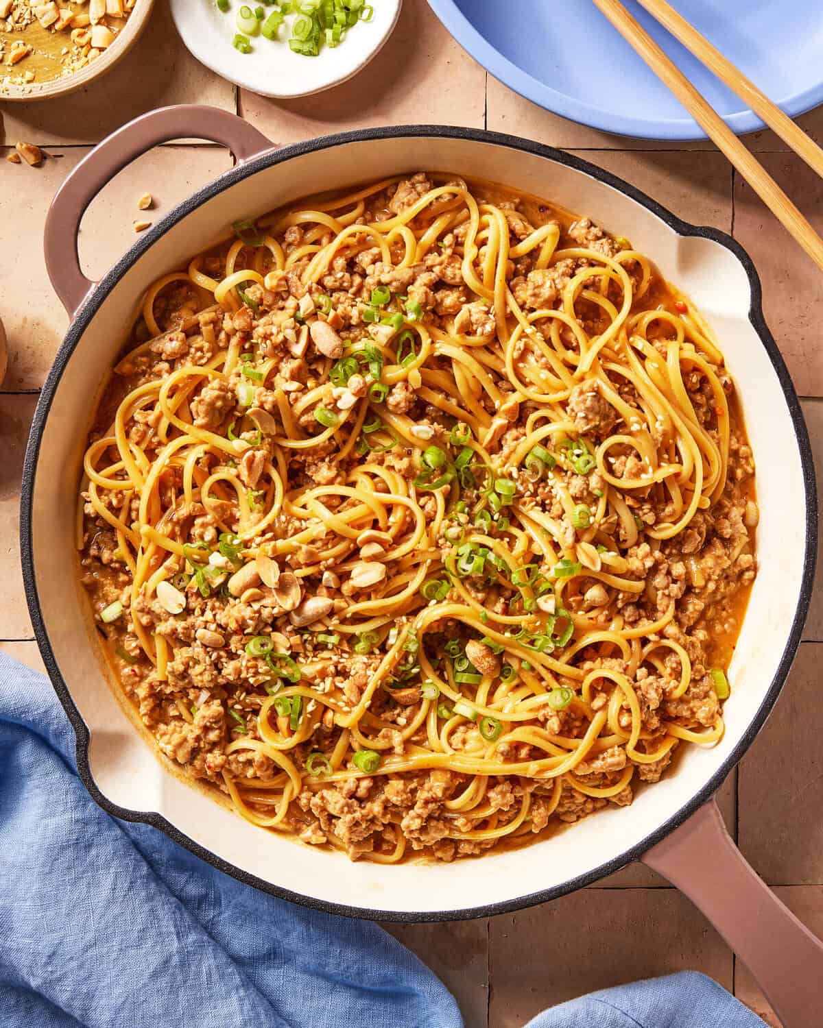 A skillet filled with noodles mixed with ground meat, topped with chopped peanuts and green onions. Chopsticks, small bowls of green onions, and a blue cloth are nearby.