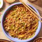 A blue bowl filled with noodles topped with ground meat, chopped green onions, and crushed peanuts, with a pair of wooden chopsticks resting on the bowl. A glass of water and bowls of sesame seeds and nuts are nearby.