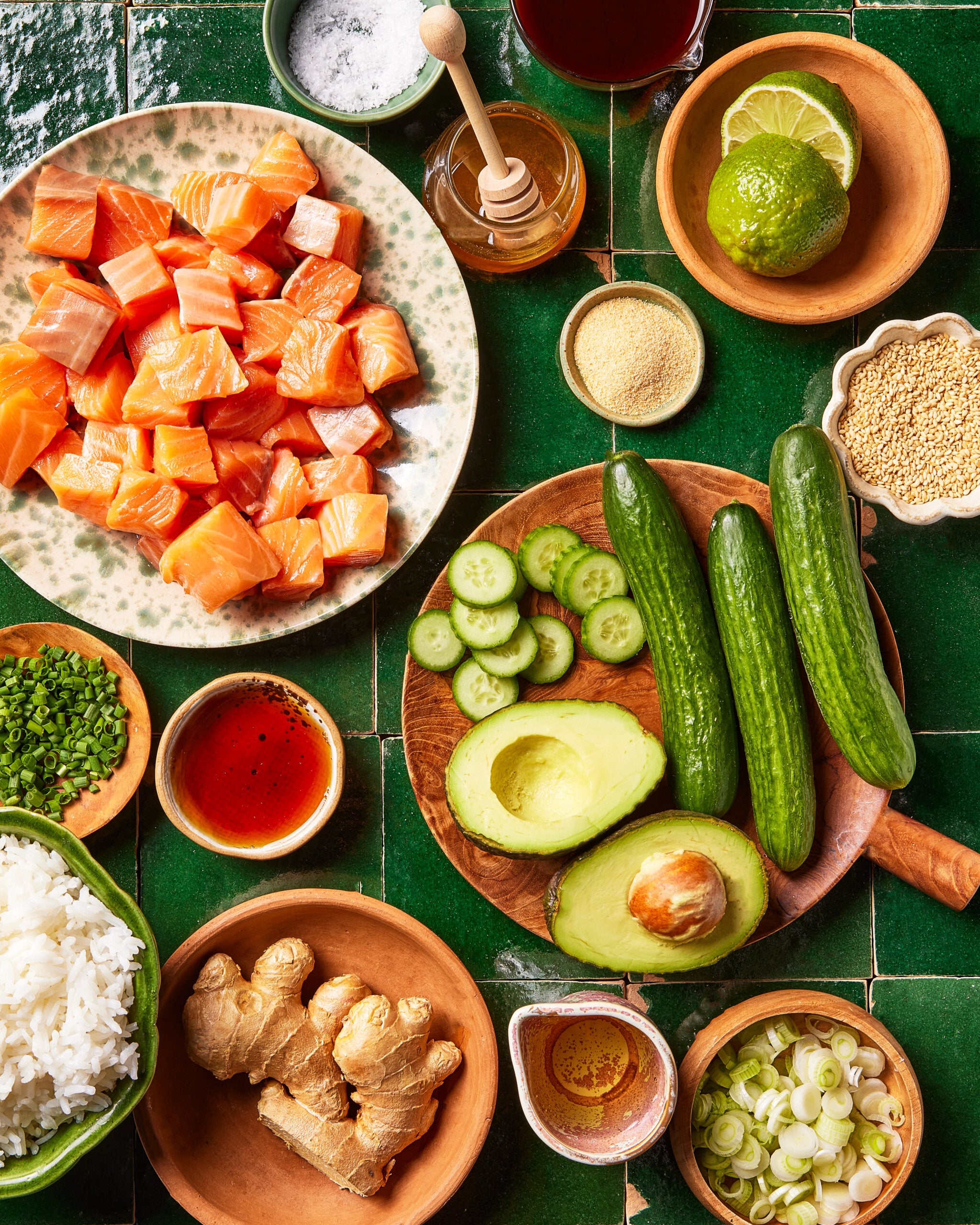 A flat lay of fresh poke bowl ingredients on a green tiled surface, including cubed raw salmon, rice, cucumbers, avocado, lime, ginger, chives, green onions, sesame seeds, honey, and various sauces in bowls.