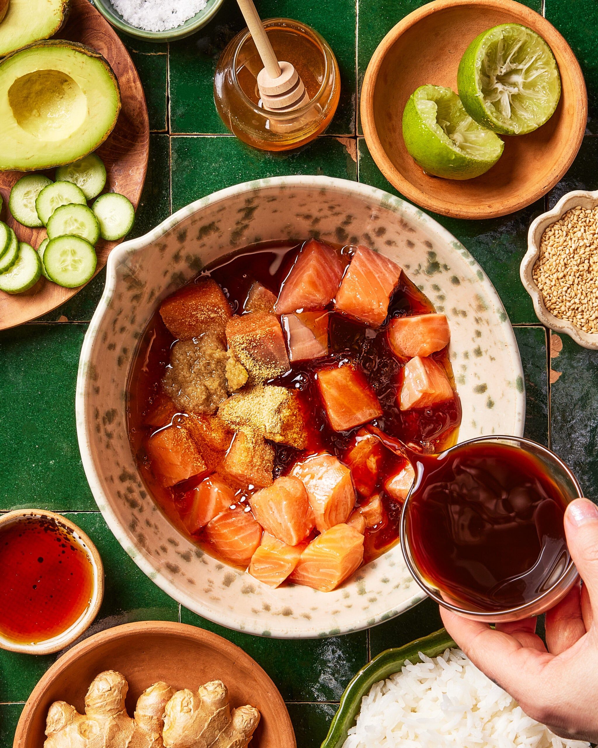 A hand pours soy sauce over cubed raw salmon in a bowl, surrounded by rice, sliced cucumber, avocado, ginger, sesame seeds, honey, a squeezed lime, and spicy sauce on a green tiled surface.