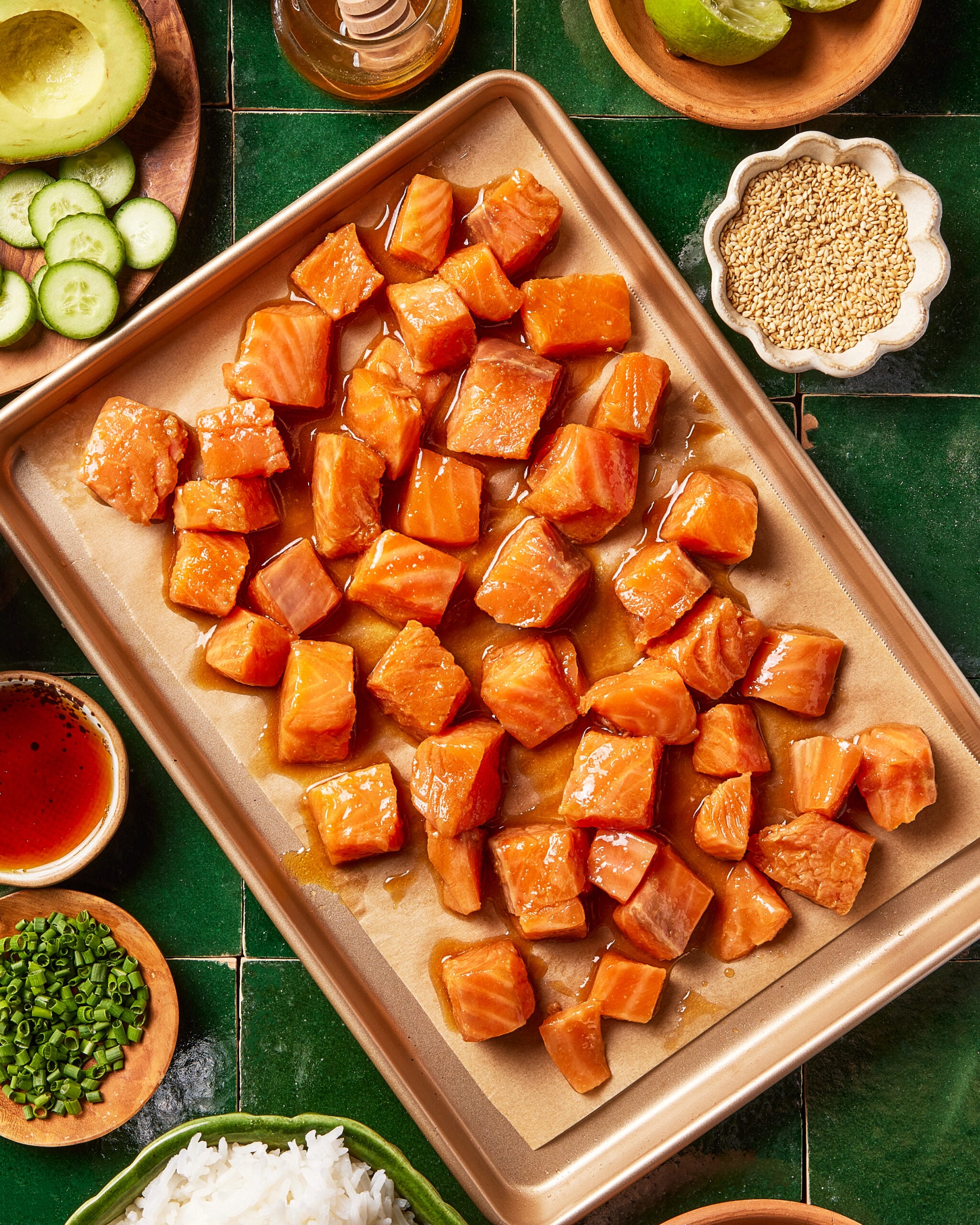A baking sheet with cubed, marinated salmon pieces surrounded by bowls of sliced cucumber, rice, chives, sauce, mustard seeds, limes, and a bottle of oil on a green tiled surface.