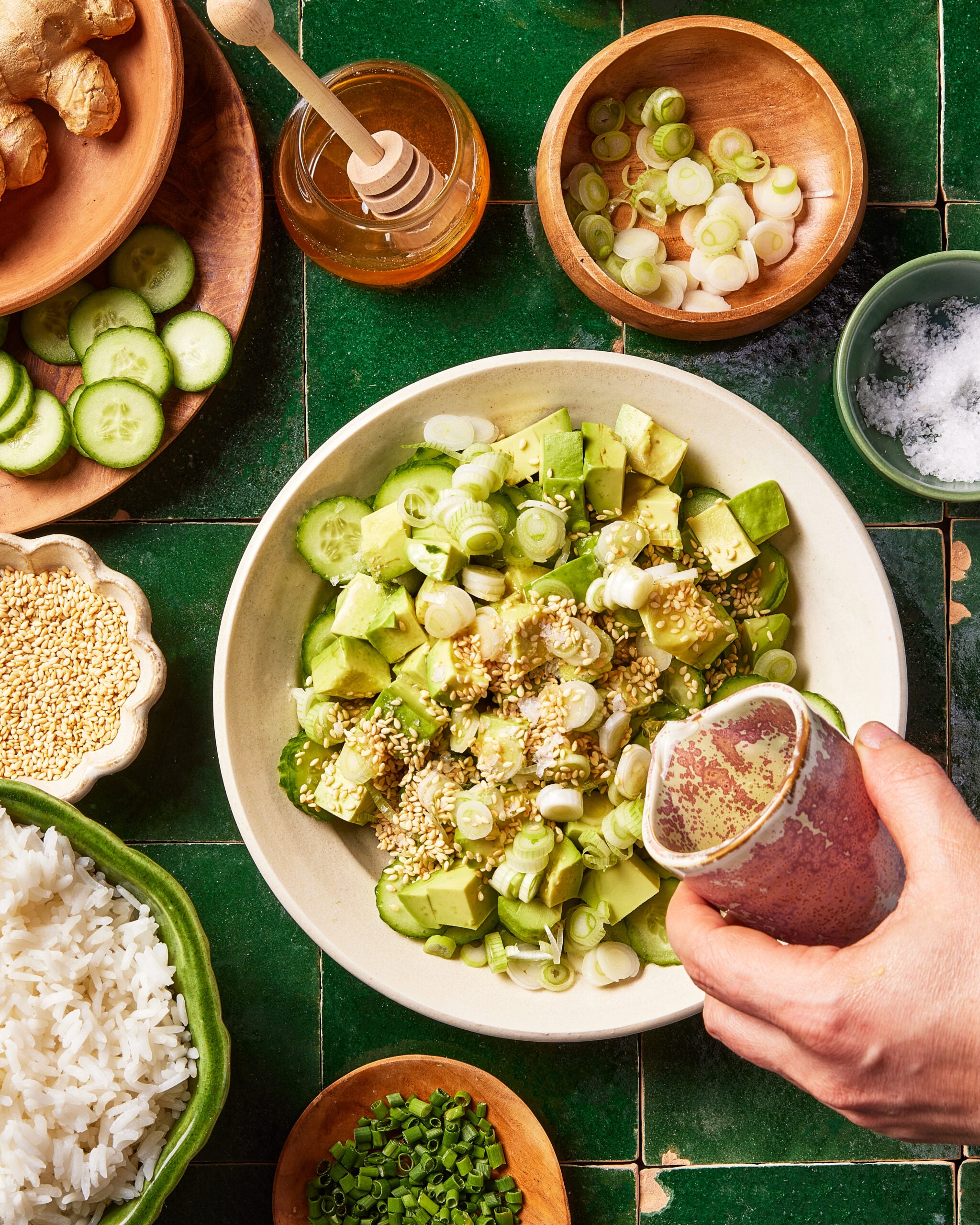 A hand pours dressing over a bowl of chopped avocado, scallions, and sesame seeds. Surrounding the bowl are rice, cucumber slices, honey, salt, ginger, and chopped chives on a green tiled surface.
