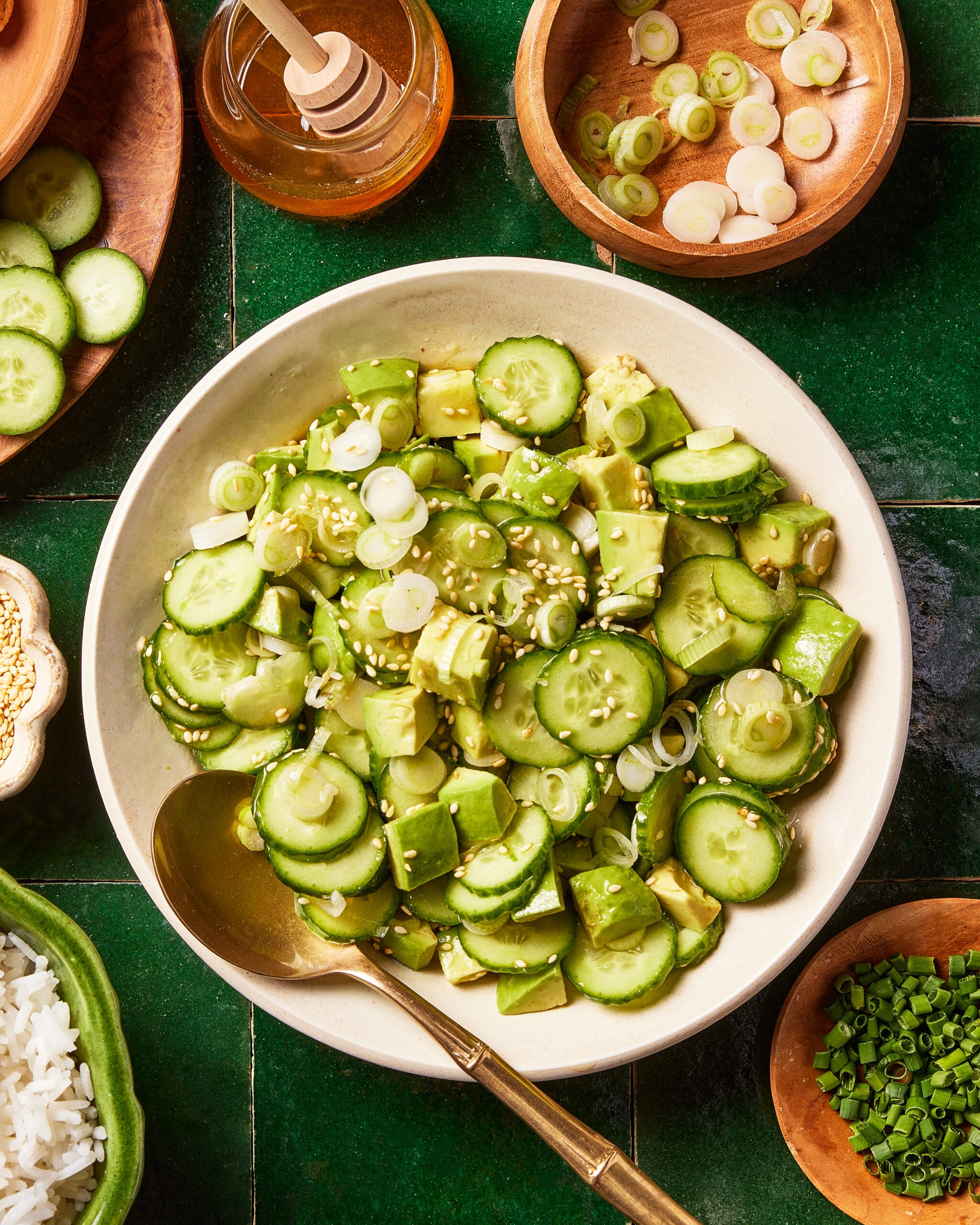 A bowl of cucumber and avocado salad topped with sliced scallions and sesame seeds sits on a green tiled surface, surrounded by bowls of rice, sliced scallions, chopped chives, and a jar of honey with a dipper.