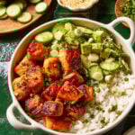 A bowl filled with white rice, glazed salmon chunks, sliced cucumbers, and chopped green onions, garnished with sesame seeds. Fresh cucumber slices and sesame seeds are visible in the background.