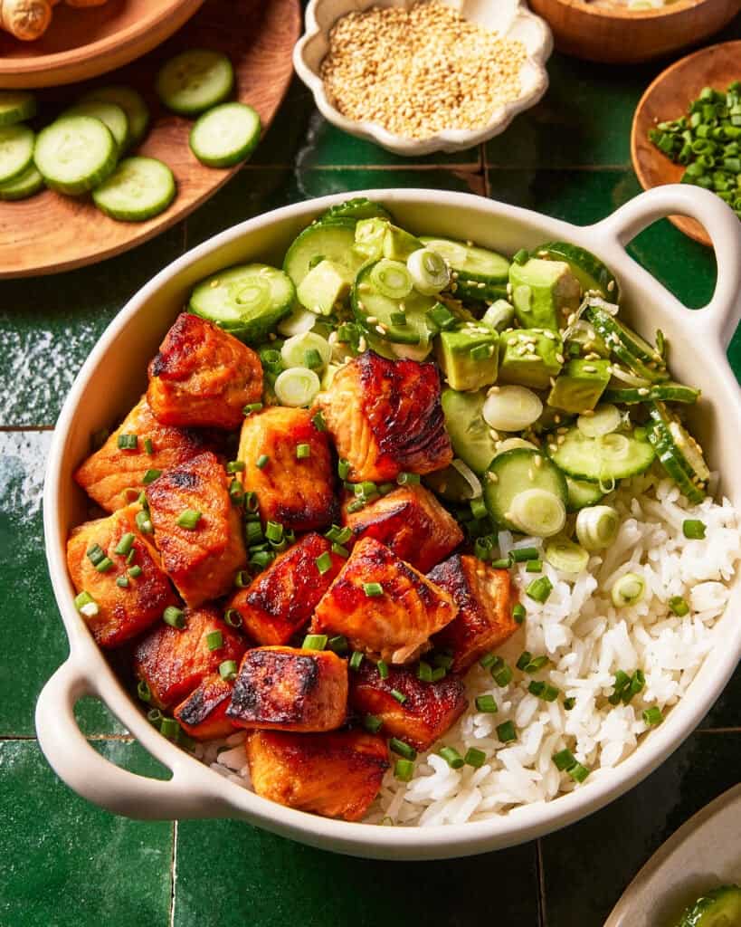 A bowl filled with white rice, glazed salmon chunks, sliced cucumbers, and chopped green onions, garnished with sesame seeds. Fresh cucumber slices and sesame seeds are visible in the background.
