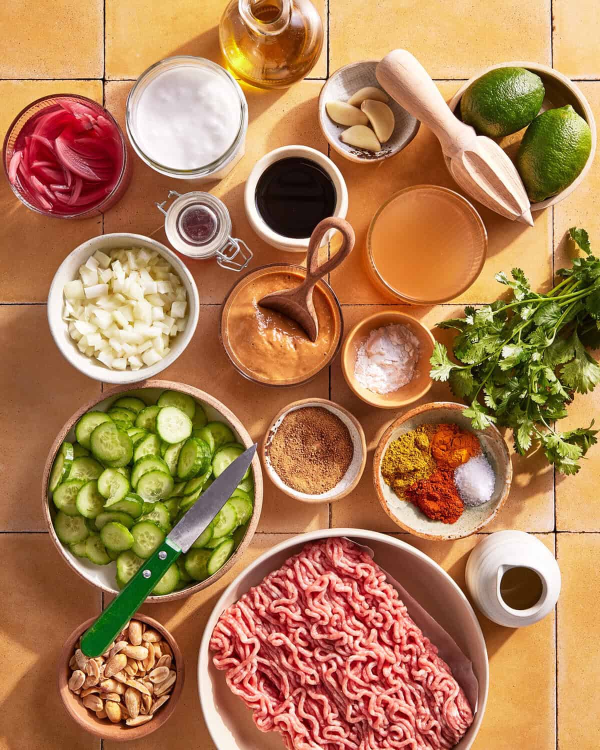 Various ingredients are neatly arranged on a tiled surface, including ground meat, sliced cucumber, onion, spices, herbs, limes, garlic, peanuts, creamy sauces, pickled onions, and bottles of oil and vinegar. A knife rests beside the cucumbers.