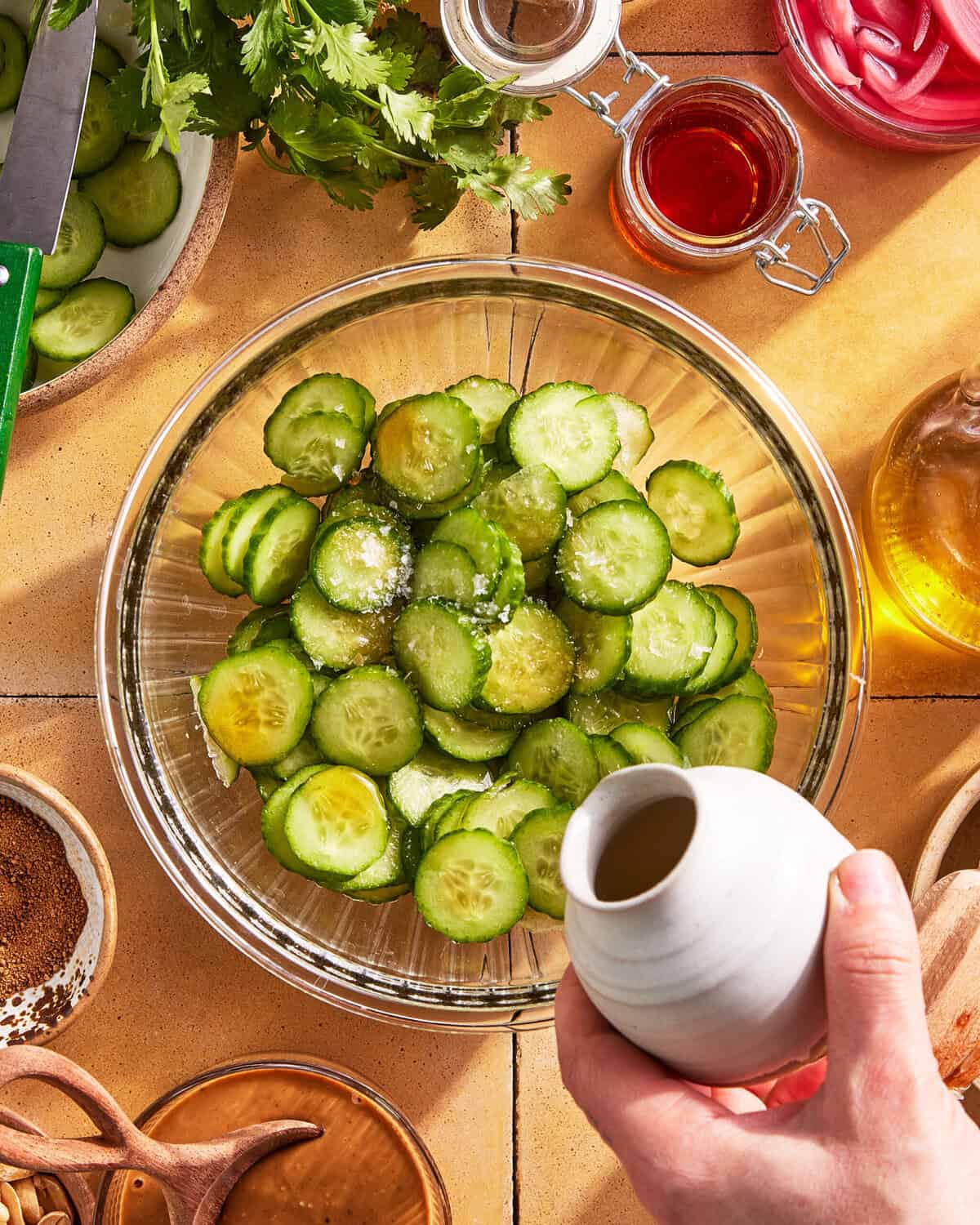 A hand pours liquid from a small white pitcher onto sliced cucumbers in a glass bowl, surrounded by herbs, seasonings, and jars on a tiled surface.