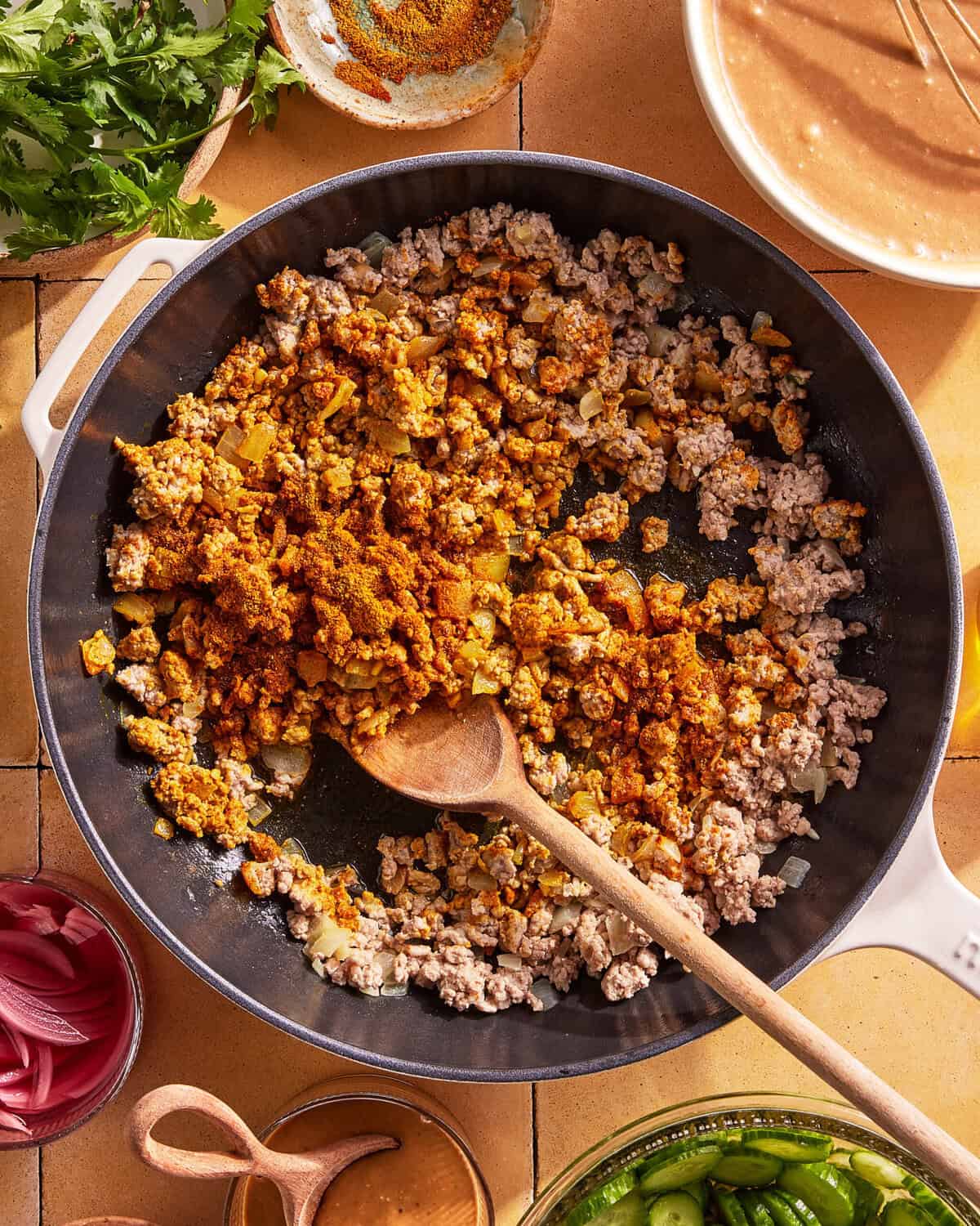 Ground meat cooking in a skillet with onions and spices being stirred with a wooden spoon. Surrounding the pan are bowls of herbs, sliced onions, cucumbers, and sauce on a tiled surface.