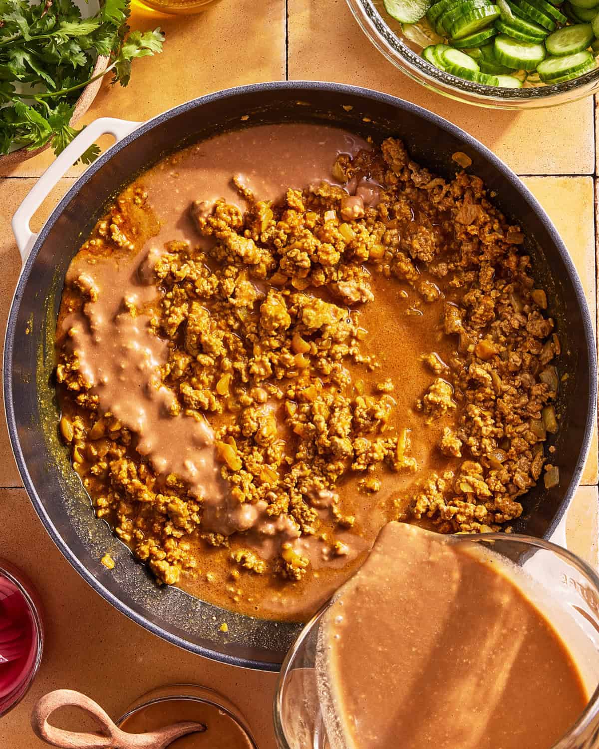 A skillet filled with cooked ground meat being mixed with a brown sauce poured from a glass container, surrounded by ingredients like sliced cucumbers, cilantro, and a bowl of red onions on a tiled surface.