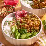 A bowl filled with white rice, sliced cucumbers, pickled red onions, chopped peanuts, and a saucy ground meat mixture, garnished with herbs. Another similar bowl and ingredients are in the background.