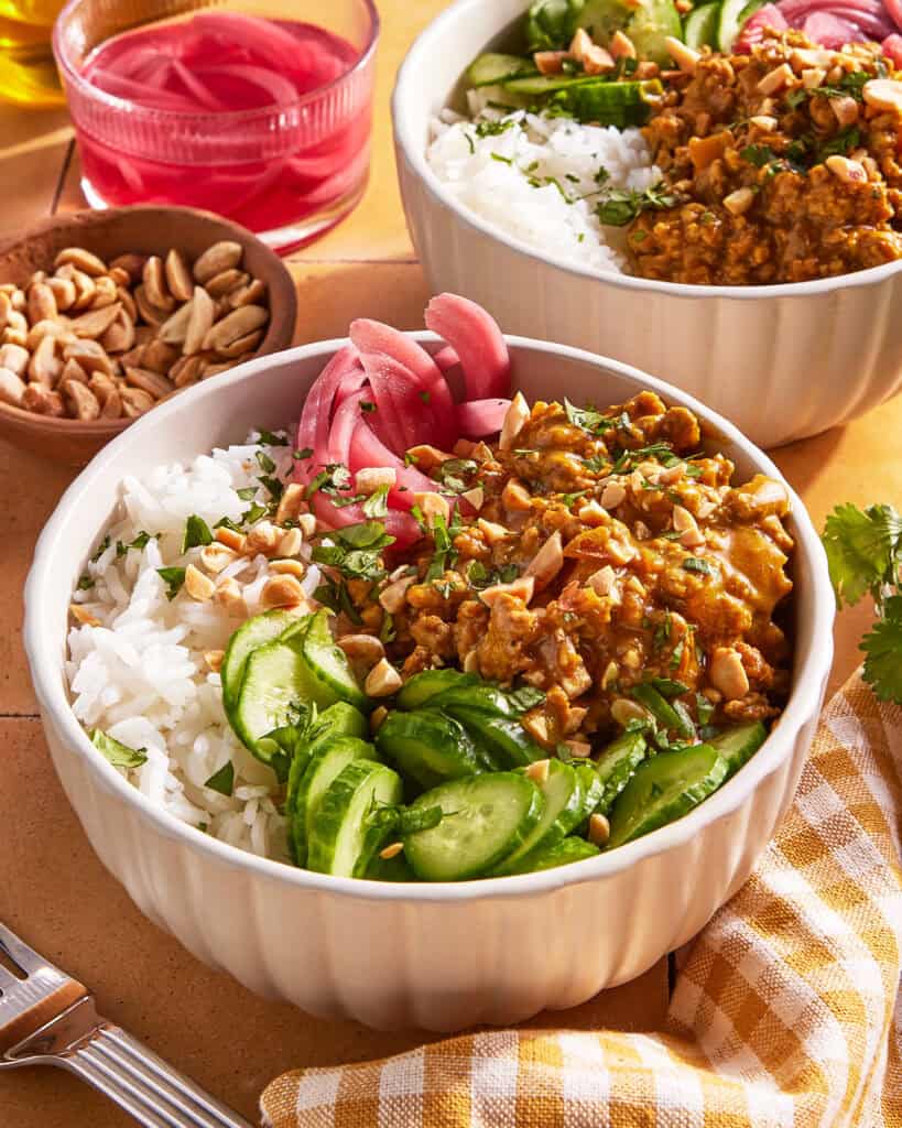 A bowl filled with white rice, sliced cucumbers, pickled red onions, chopped peanuts, and a saucy ground meat mixture, garnished with herbs. Another similar bowl and ingredients are in the background.