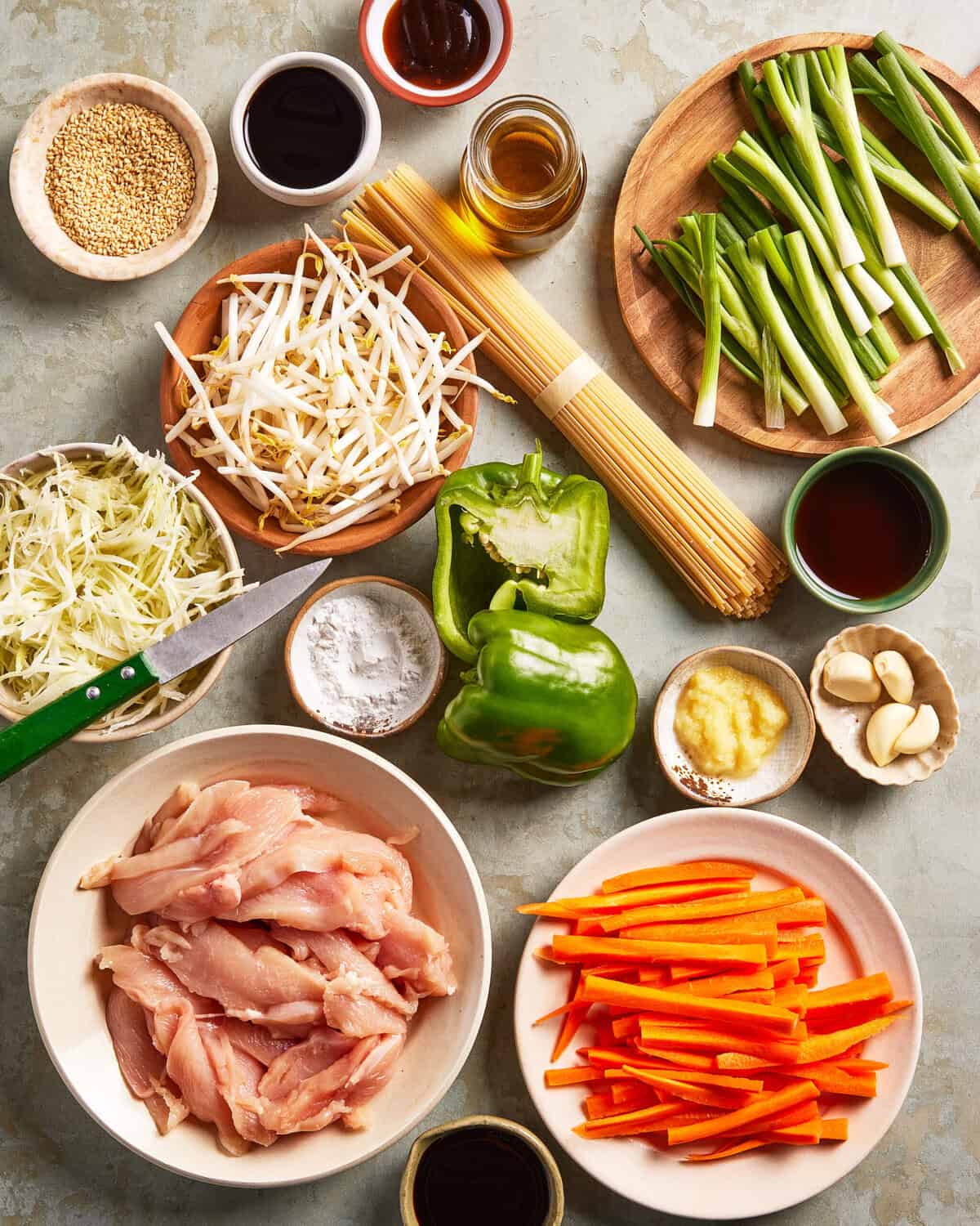 A top view of various fresh stir-fry ingredients arranged on a table, including sliced chicken, carrots, green bell pepper, cabbage, bean sprouts, green onions, garlic, sauces, sesame seeds, spaghetti, ginger, and seasonings.