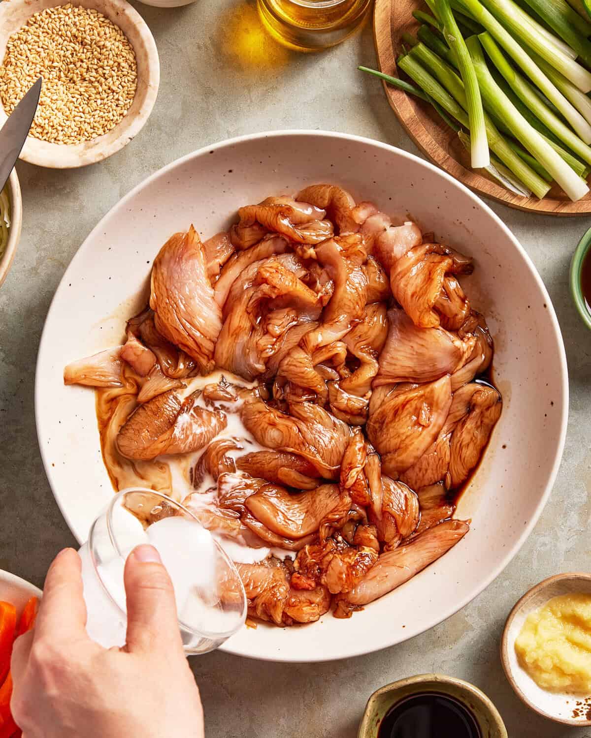 A hand pours a clear liquid into a bowl of marinated raw chicken. Surrounding the bowl are sliced green onions, sesame seeds, oil, and dipping sauces on a light countertop.