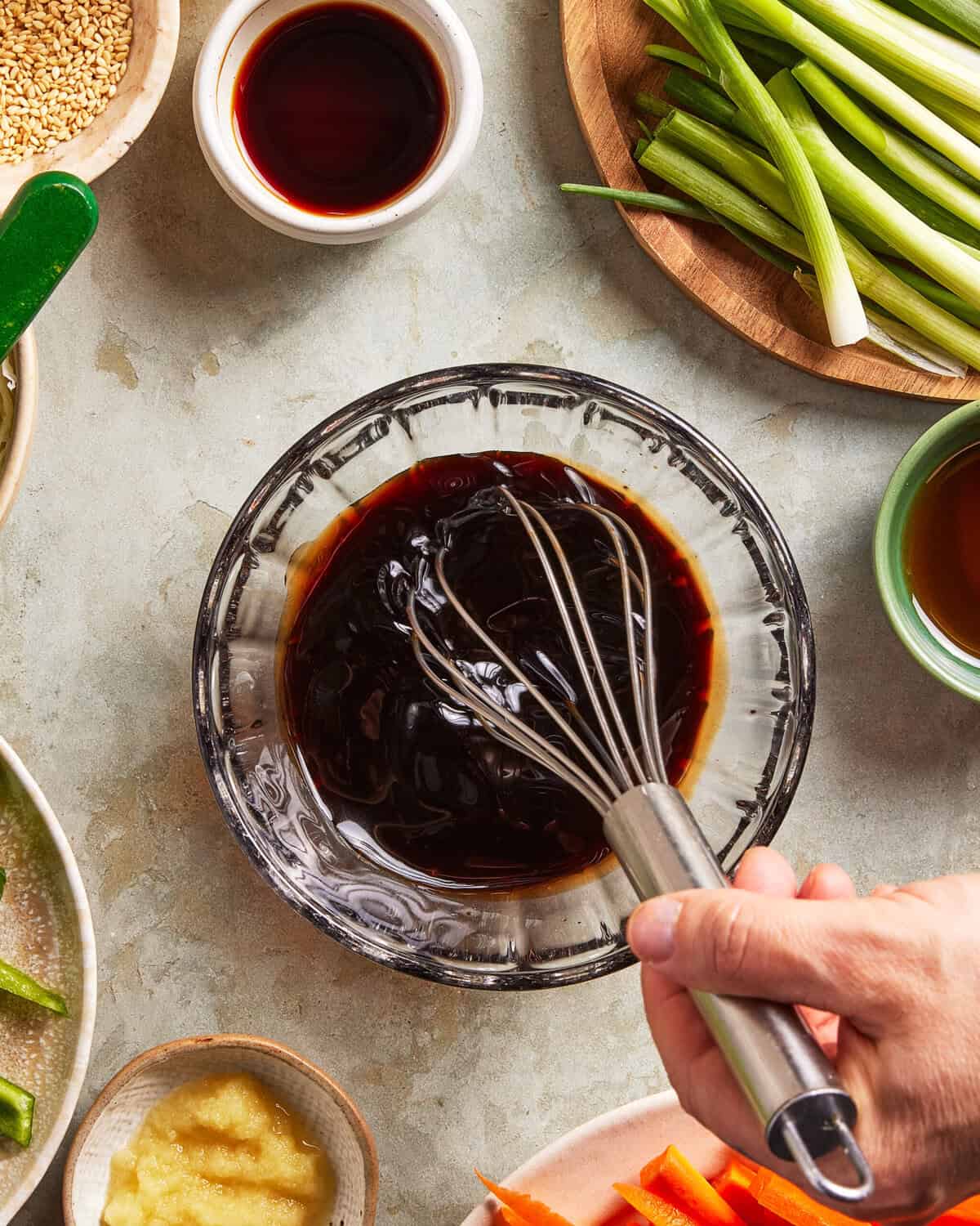 A hand whisks a dark sauce in a glass bowl. Surrounding the bowl are ingredients such as sliced green onions, soy sauce, sesame seeds, ginger paste, and carrot sticks on a light-colored surface.