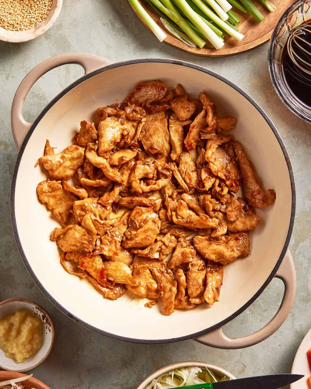 A large beige pot filled with cooked, sliced pieces of seasoned chicken sits on a countertop surrounded by small bowls containing green onions, ginger, sesame seeds, and dark sauce.