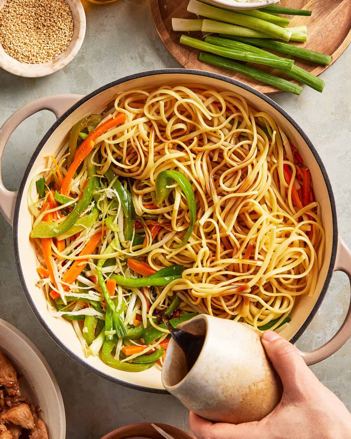 A hand pours soy sauce into a large pot containing cooked noodles, sliced green and orange bell peppers, carrots, and green onions. Bowls of sesame seeds, green onions, and other ingredients are nearby.