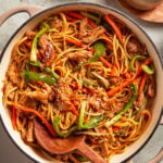 A pot filled with stir-fried noodles, sliced chicken, bell peppers, carrots, and green onions, garnished with sesame seeds. A wooden spoon rests inside the pot, and a bowl of sesame seeds sits nearby.