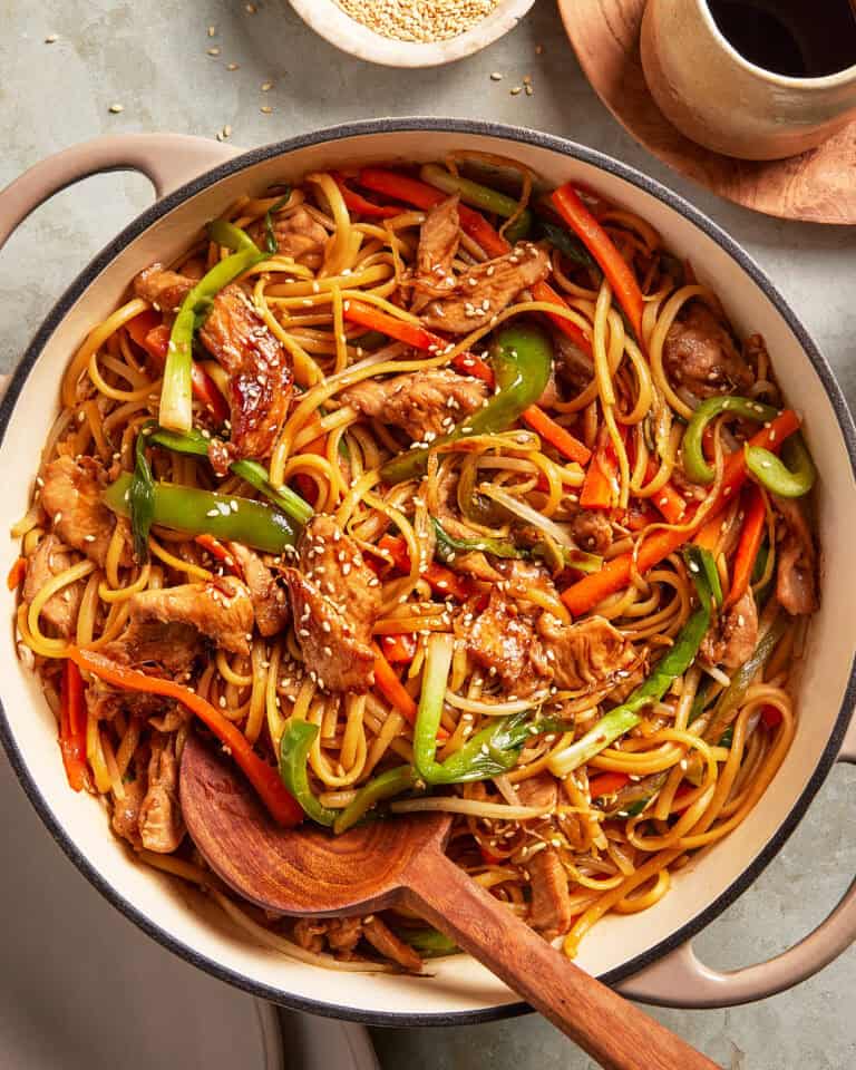 A pot filled with stir-fried noodles, sliced chicken, bell peppers, carrots, and green onions, garnished with sesame seeds. A wooden spoon rests inside the pot, and a bowl of sesame seeds sits nearby.