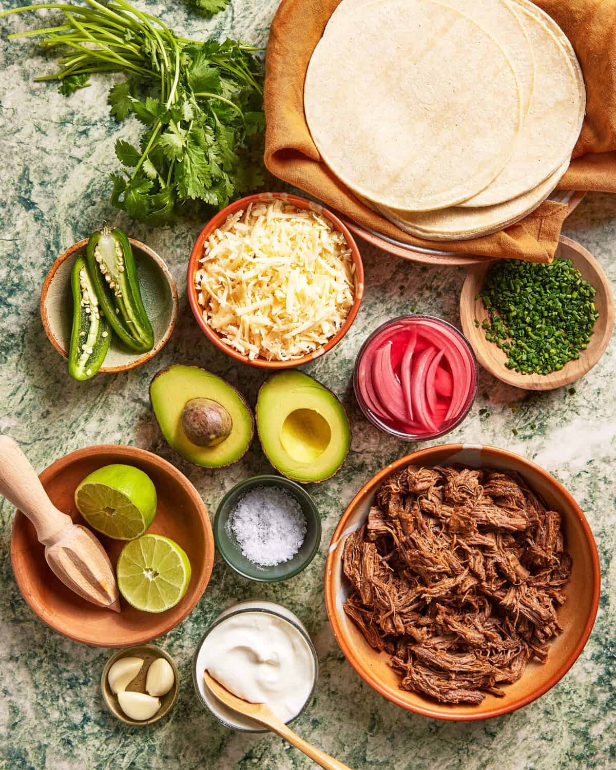 An overhead view of taco ingredients on a green marble surface: shredded beef, corn tortillas, avocado halves, lime, garlic, sliced cheese, chopped herbs, sliced jalapeños, pickled onions, sour cream, salt, and fresh cilantro.