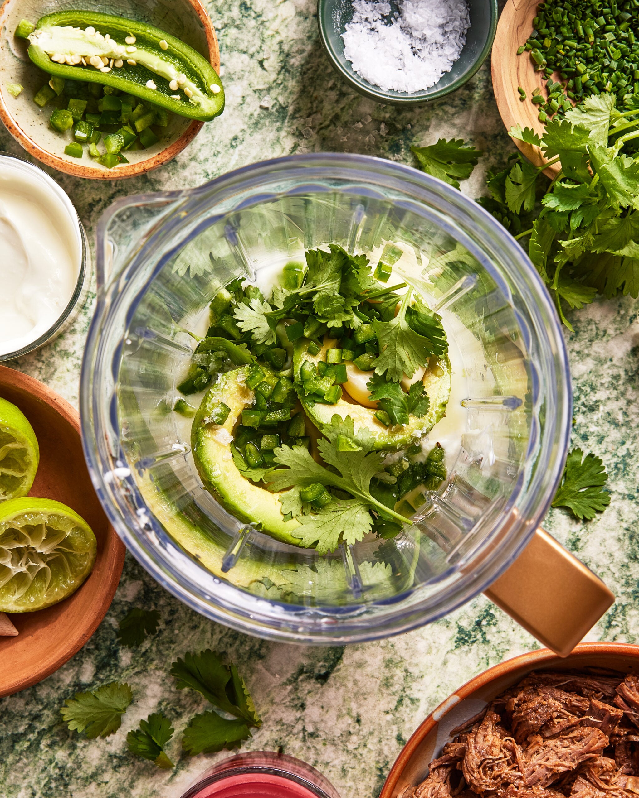 A blender filled with avocado, chopped jalapeño, and cilantro sits on a counter surrounded by bowls of lime, sour cream, shredded meat, salt, fresh herbs, and other ingredients.