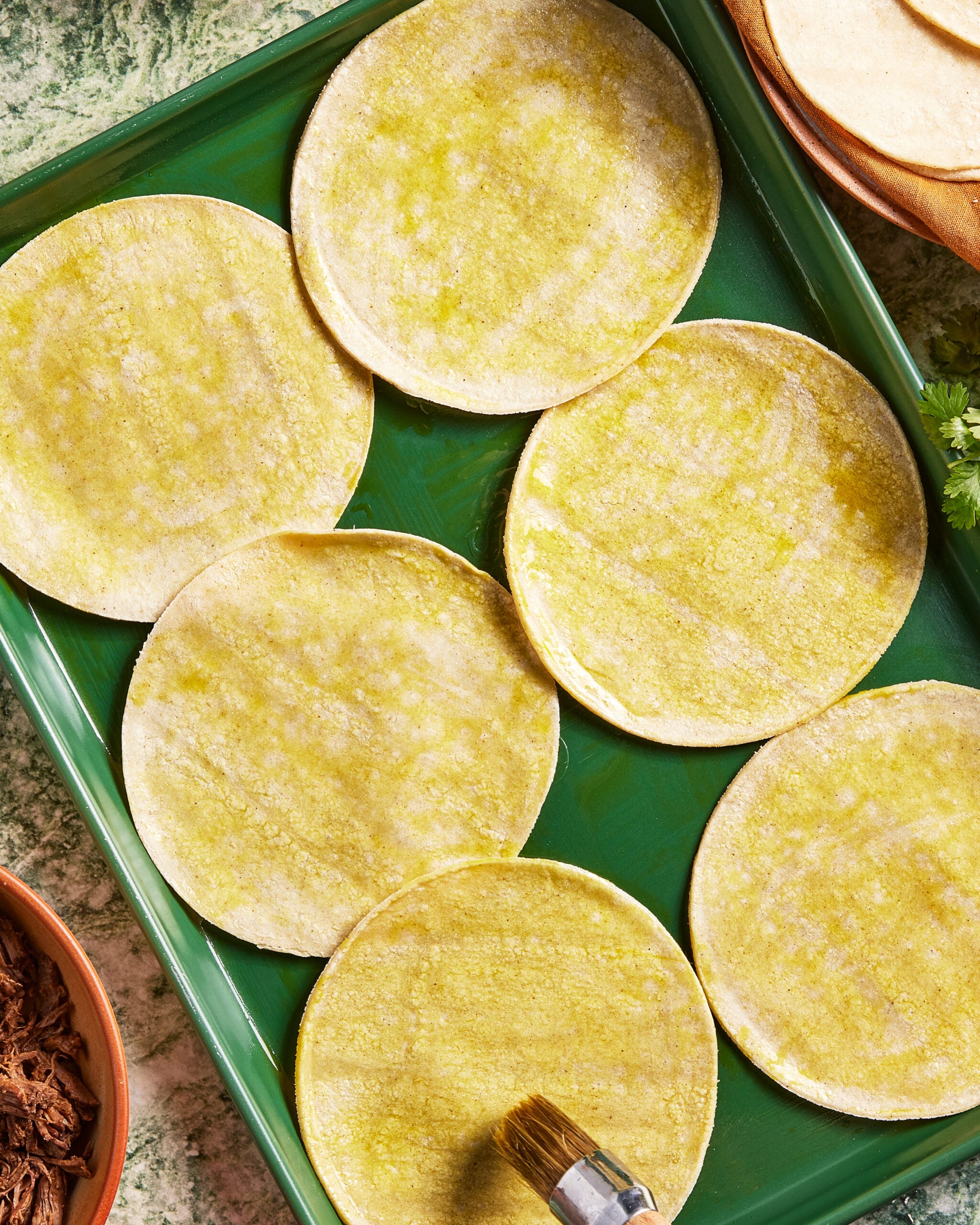 Six corn tortillas are laid out on a green tray, brushed with oil. A small brush is visible in the foreground. The scene is set on a green marble surface, with some shredded meat and fresh cilantro visible at the edges.