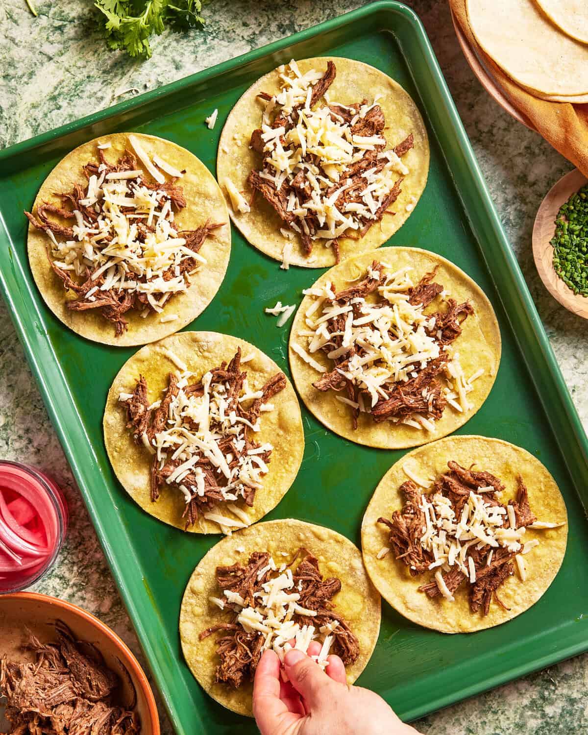 A green baking sheet holds six corn tortillas topped with shredded beef and cheese. A hand is adding cheese to one tortilla. Surrounding the tray are bowls of ingredients and stacks of tortillas.