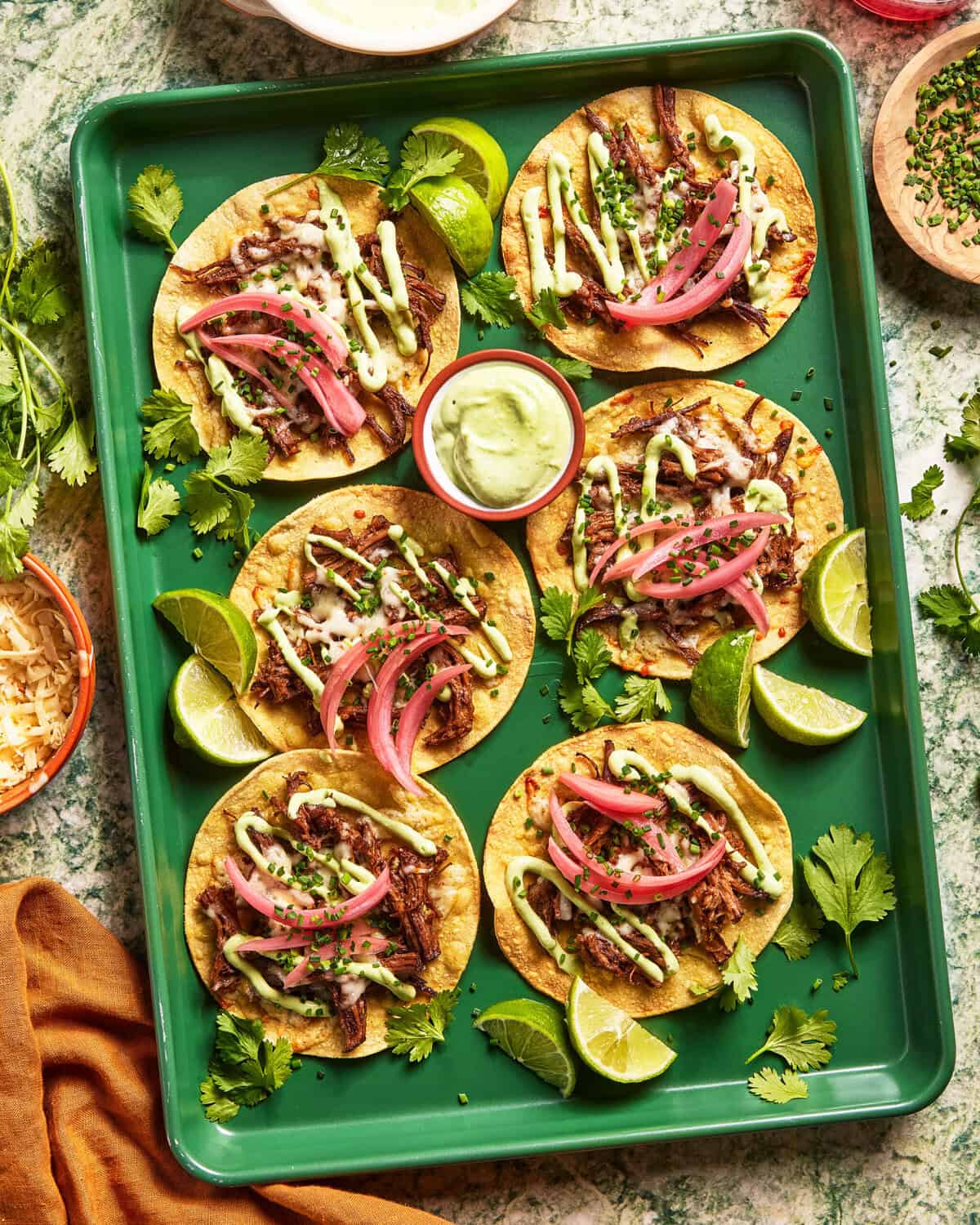 A green tray with six tacos topped with shredded meat, pickled red onions, and green sauce, garnished with fresh cilantro and lime wedges. A small bowl of creamy green sauce is in the center of the tray.