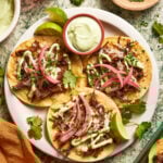 Three shredded beef tostadas topped with melted cheese, pickled red onions, avocado crema, and chopped chives. Served on a plate with lime wedges, tortilla chips, and a small bowl of avocado sauce.