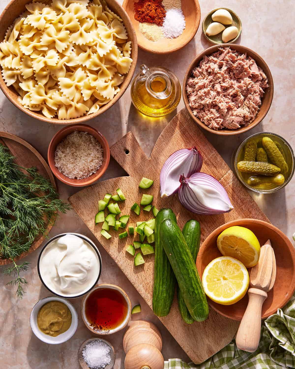 A top-down view of ingredients for a pasta salad, including bowtie pasta, canned tuna, cucumbers, red onion, lemon, mayo, mustard, oil, pickles, garlic, dill, breadcrumbs, spices, salt, and pepper on a cutting board.