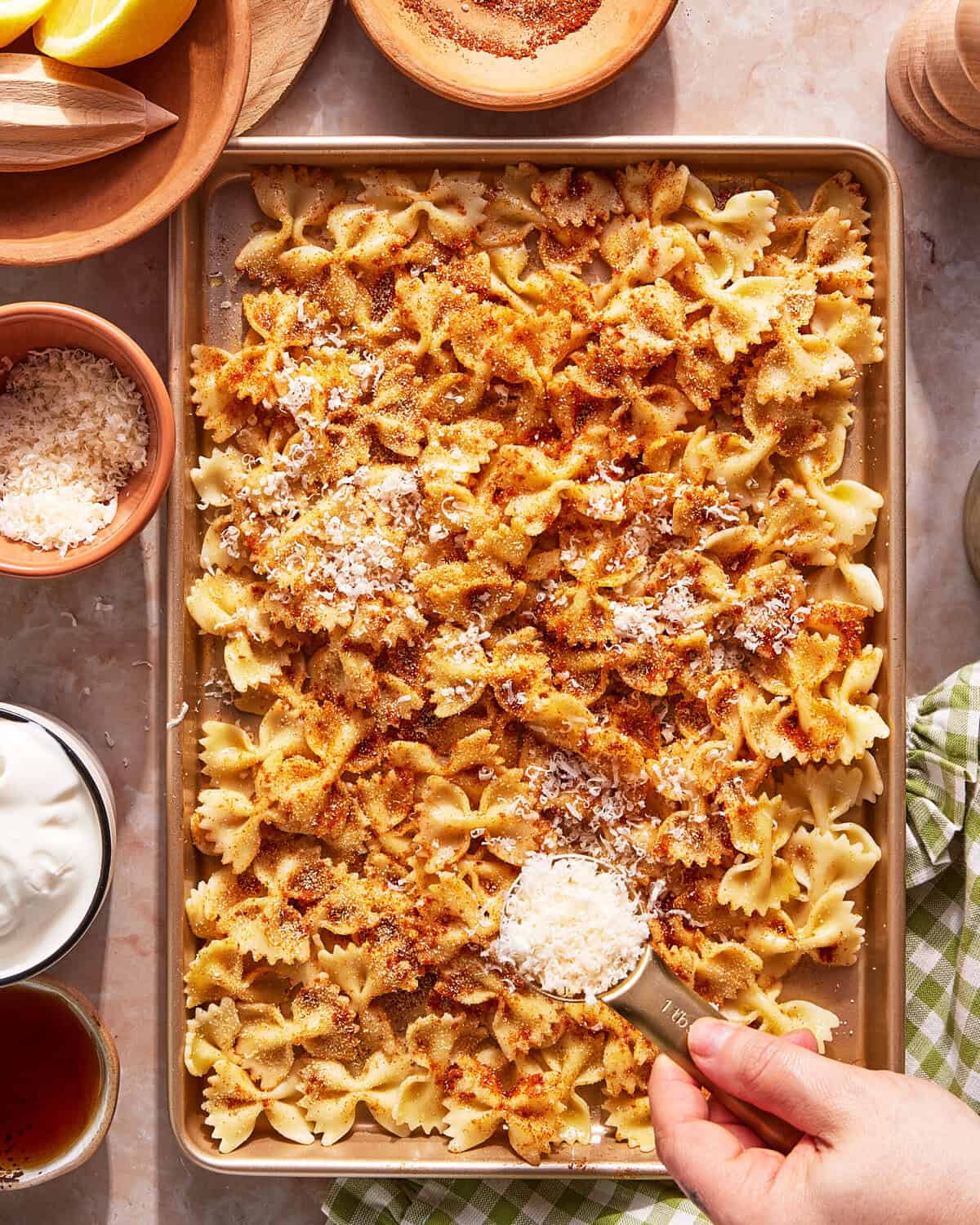 A hand sprinkles grated cheese over a baking sheet filled with baked bowtie pasta topped with golden breadcrumbs. Small bowls of ingredients and a green-checked napkin are nearby on a marble surface.