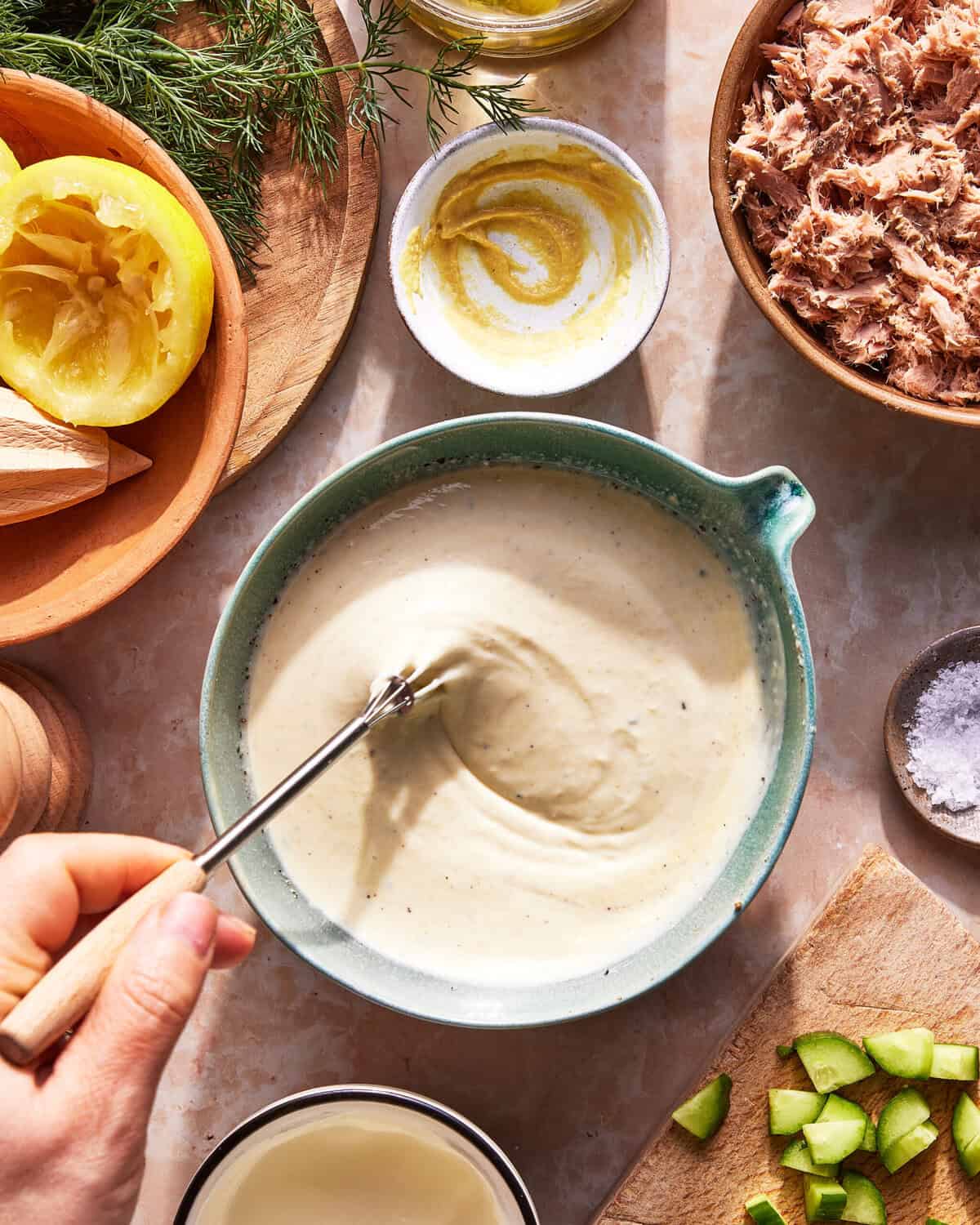 A hand whisks a creamy sauce in a bowl, surrounded by ingredients including lemon, fresh dill, tuna, mustard, salt, and chopped celery on a kitchen counter.