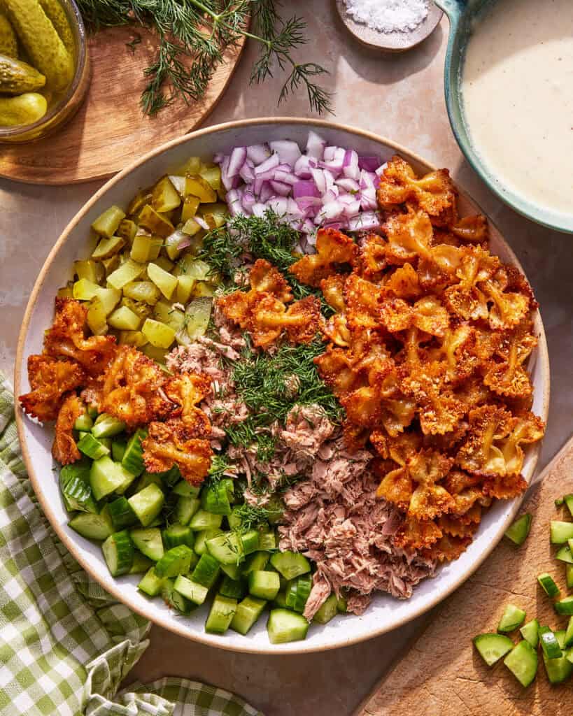 A bowl filled with crispy bowtie pasta, chopped pickles, diced red onion, fresh dill, tuna, and cucumber, arranged in sections on a light surface with a bowl of dressing and herbs nearby.