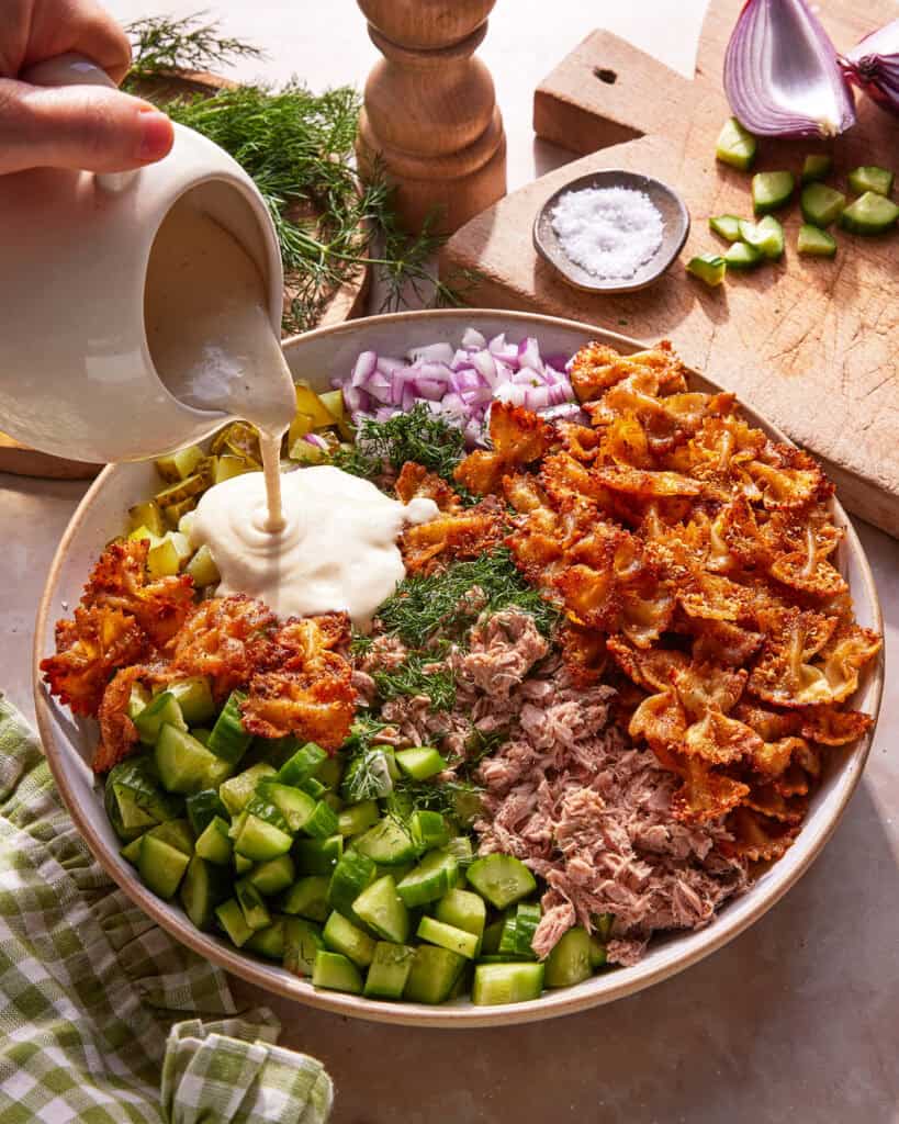 A bowl with cooked bowtie pasta, tuna, chopped cucumber, red onion, fresh dill, and creamy dressing being poured, surrounded by fresh ingredients and a wooden pepper grinder on a kitchen counter.